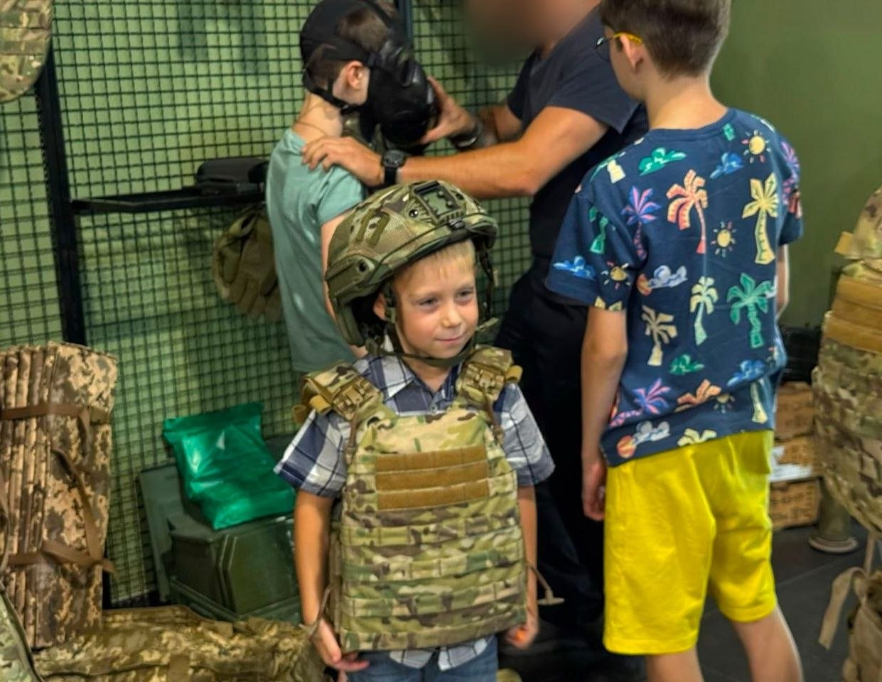 Хлопчик молодшого віку з-поміж переселенців із Херсонщини приміряє військову амуніцію у відкритому просторі «Артан» в Одесі. Дитина в картатій сорочці стоїть по центру, вдягнена у великий камуфляжний бронежилет та захисний шолом. Праворуч спиною до камери стоїть інший хлопчик у жовтих шортах і кольоровій футболці. На задньому плані чоловік, чиє обличчя розмите з міркувань безпеки, допомагає третій дитині одягнути протигаз. Навколо них видно елементи військового спорядження, маскувальні сітки та ящики.