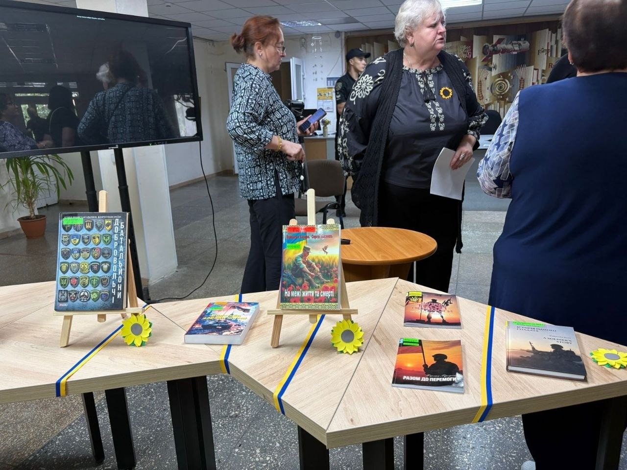 Книжкова виставка-меморіал зблизька під час патріотичного заходу для переселенців у Кропивницькому. На дерев'яних столах, з'єднаних півколом і прикрашених жовто-блакитними стрічками та штучними соняшниками, розставлені книги про війну та українських захисників. Серед видань на маленьких дерев'яних мольбертах-підставках видно книги «Добровольчі батальйони» та «На межі життя та смерті». Ліворуч стоїть великий вимкнений телевізійний екран. На задньому плані спілкуються організатори та гості заходу, зокрема літня ведуча у вишиванці.