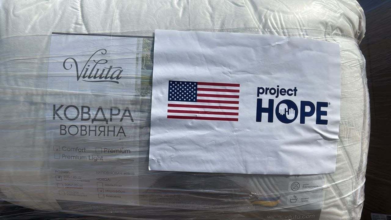 Велика вовняна ковдра білого кольору у прозорій захисній упаковці з партії гуманітарного вантажу для медичних закладів Херсонщини від благодійників. Ліворуч під плівкою видно фірмову етикетку українського виробника з написом «Viluta КОВДРА ВОВНЯНА». Праворуч поверх упаковки наклеєно великий білий аркуш із прапором США та логотипом міжнародної неурядової організації «project HOPE». Увесь пакунок із теплою ковдрою щільно обмотаний прозорою транспортувальною стрейч-плівкою.