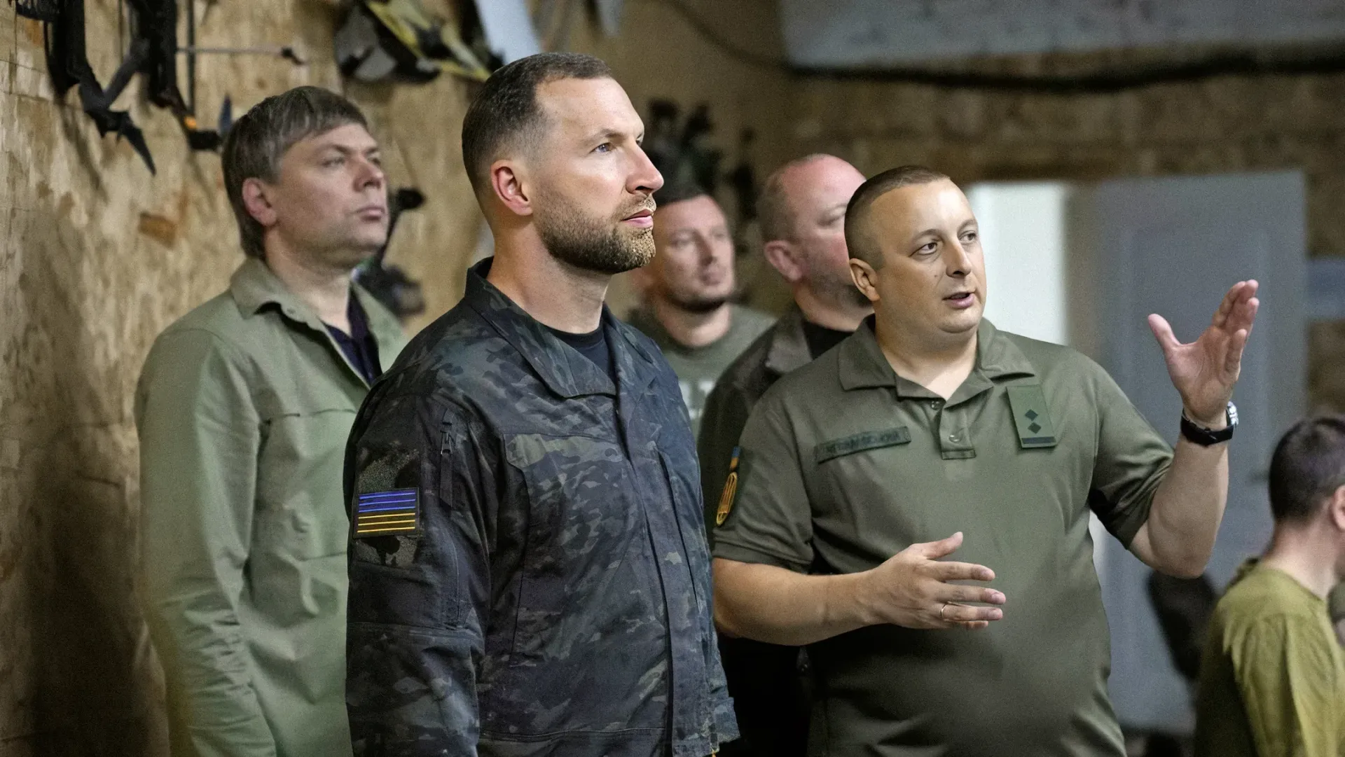 Робоча поїздка Міністра оборони України на Херсонщину. На передньому плані по центру стоїть Міністр оборони Євгеній Хмара у чорній тактичній сорочці з прапором України на рукаві. Праворуч від нього — військовий у футболці кольору хакі з шевроном на рукаві, який активно доповідає, жестикулюючи піднятою правою рукою. На задньому плані за плечем Міністра видно начальника Херсонської ОВА Олександра Прокудіна, який уважно слухає разом із рештою присутніх військових та посадовців. Зустріч проходить у закритому приміщенні з дерев'яним оздобленням стін. У лівому верхньому кутку фото розміщено білий логотип Міністерства оборони України у формі тризуба на темному щиті.