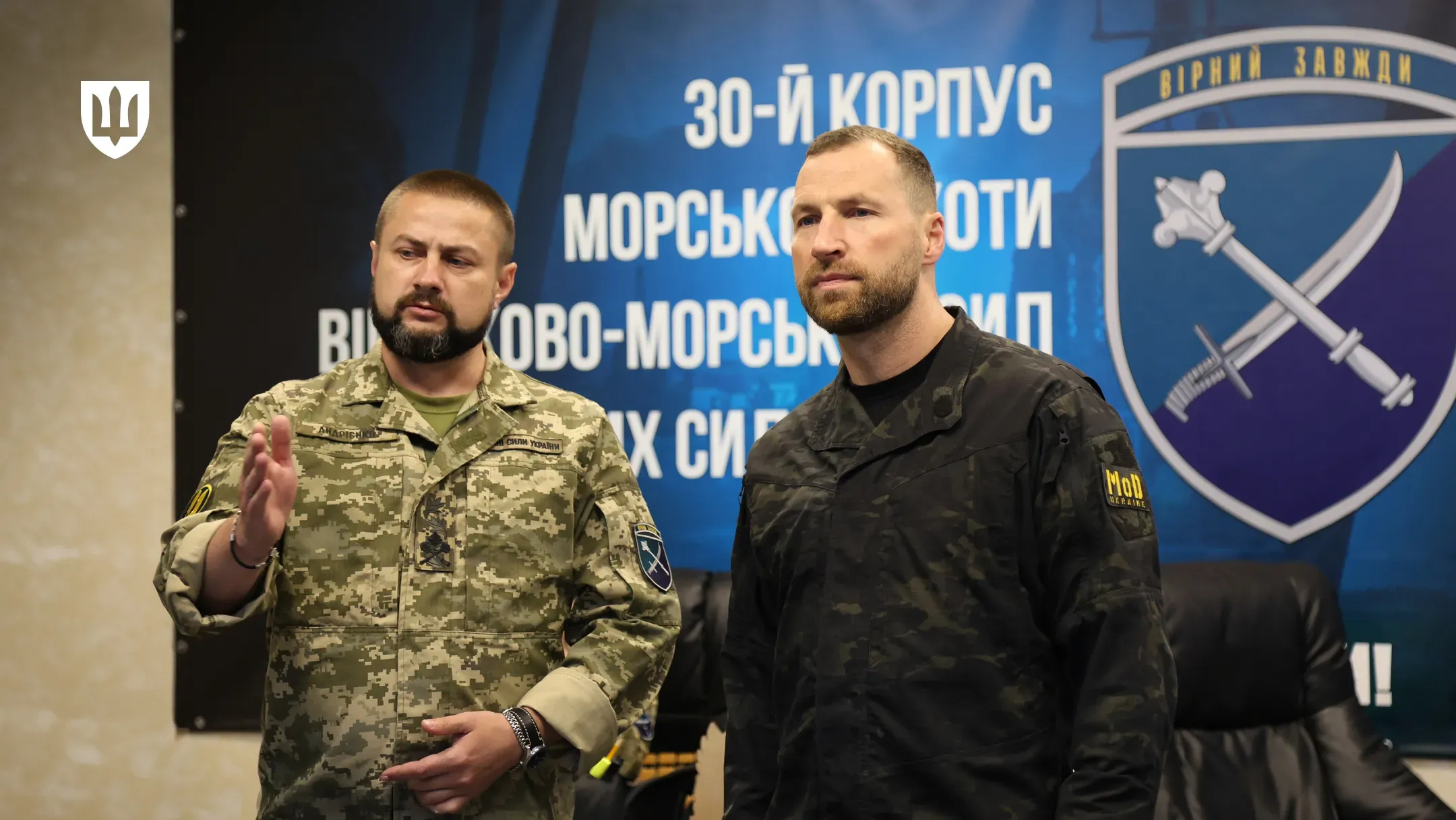 Робоча поїздка Міністра оборони України на Херсонщину. У приміщенні на тлі великого синього банера з написом «30-Й КОРПУС МОРСЬКОЇ ПІХОТИ» стоять двоє чоловіків. Праворуч — Міністр оборони Євгеній Хмара у чорній тактичній сорочці з шевроном «MoD» (Міністерство оборони) на рукаві, який уважно слухає співрозмовника. Ліворуч від нього — військовий у камуфляжній формі ЗСУ, який активно щось пояснює, піднявши праву руку в жесті. У лівому верхньому кутку кадру розміщено білий логотип Міністерства оборони України у формі тризуба на темному щиті.