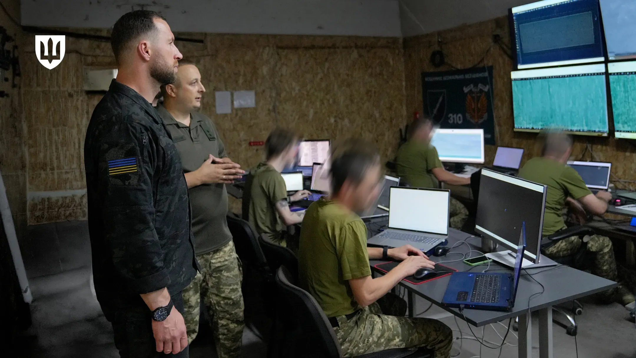 Робоча поїздка Міністра оборони України на Херсонщину. Міністр оборони Євгеній Хмара у чорній тактичній сорочці з прапором України на рукаві стоїть у командному пункті чи центрі управління. Поруч із ним стоїть військовий у футболці кольору хакі та камуфляжних штанах, який зводить долоні разом під час розмови. Вони спостерігають за роботою операторів. У залі розсаджені військовослужбовці за робочими місцями з ноутбуками та великими моніторами, на яких відображаються дані та графіки (обличчя та екрани розмиті з міркувань безпеки). На стіні на задньому плані видно темний прапор із номером «310». У лівому верхньому кутку кадру розміщено білий логотип Міністерства оборони України у формі тризуба на темному щиті.