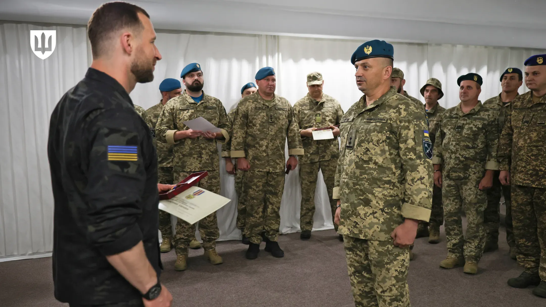 Робоча поїздка Міністра оборони України на Херсонщину. На передньому плані по центру стоїть Міністр оборони Євгеній Хмара у чорній тактичній сорочці з прапором України на рукаві. Праворуч від нього — військовослужбовець у футболці кольору хакі з шевроном Сухопутних військ (або ЗСУ) на рукаві, який активно доповідає, жестикулюючи піднятою правою рукою. На задньому плані та ліворуч стоять інші військовослужбовці, які уважно слухають доповідача. Зустріч проходить у закритому приміщенні з дерев'яним оздобленням стін, на яких закріплені елементи військового спорядження. У лівому верхньому кутку кадру присутній білий логотип Міністерства оборони України у формі тризуба на темному щиті.