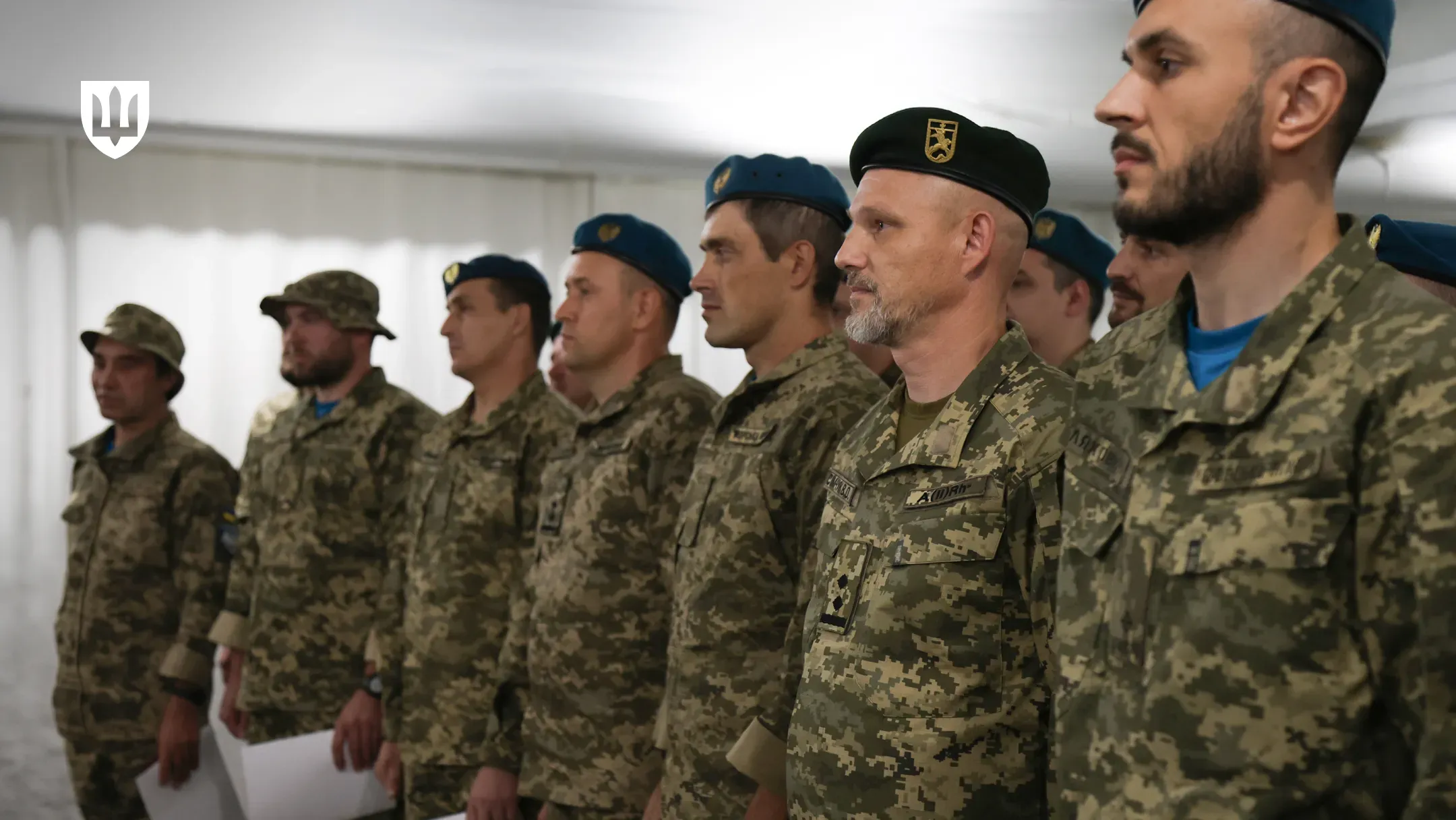 Робоча поїздка Міністра оборони України на Херсонщину. Шеренга українських військовослужбовців у камуфляжній формі ЗСУ стоїть у приміщенні під час офіційного заходу чи нагородження. На передньому плані праворуч — військові у беретах, серед яких офіцер у чорному береті з емблемою Морської піхоти та військовий у блакитному береті. Далі в ряду стоять воїни у камуфляжних панамах, деякі з них тримають у руках аркуші документів або нагородні листи. У лівому верхньому кутку кадру присутній білий логотип Міністерства оборони України у формі тризуба на темному щиті.