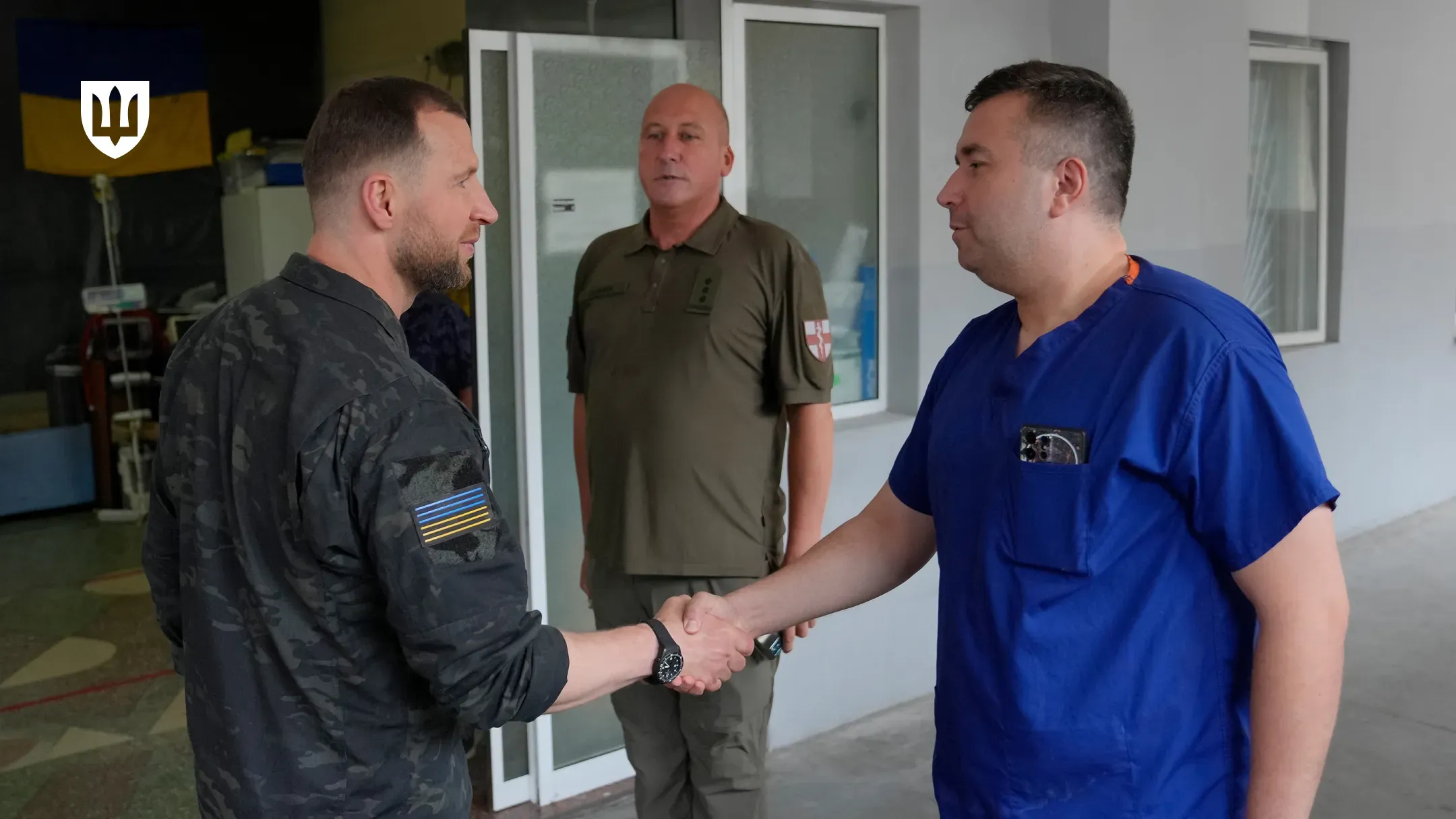Робоча поїздка Міністра оборони України на Херсонщину. Ліворуч на передньому плані — Міністр оборони Євгеній Хмара у чорній тактичній сорочці з прапором України на рукаві, який тисне руку лікарю у синьому медичному костюмі. На задньому плані по центру стоїть чоловік у військовій сорочці поло кольору хакі з шевронами на рукаві й погонами капітана, який спостерігає за зустріччю. Подія відбувається біля входу до медичного закладу. У лівому верхньому кутку кадру присутній білий логотип Міністерства оборони України у формі тризуба на темному щиті.