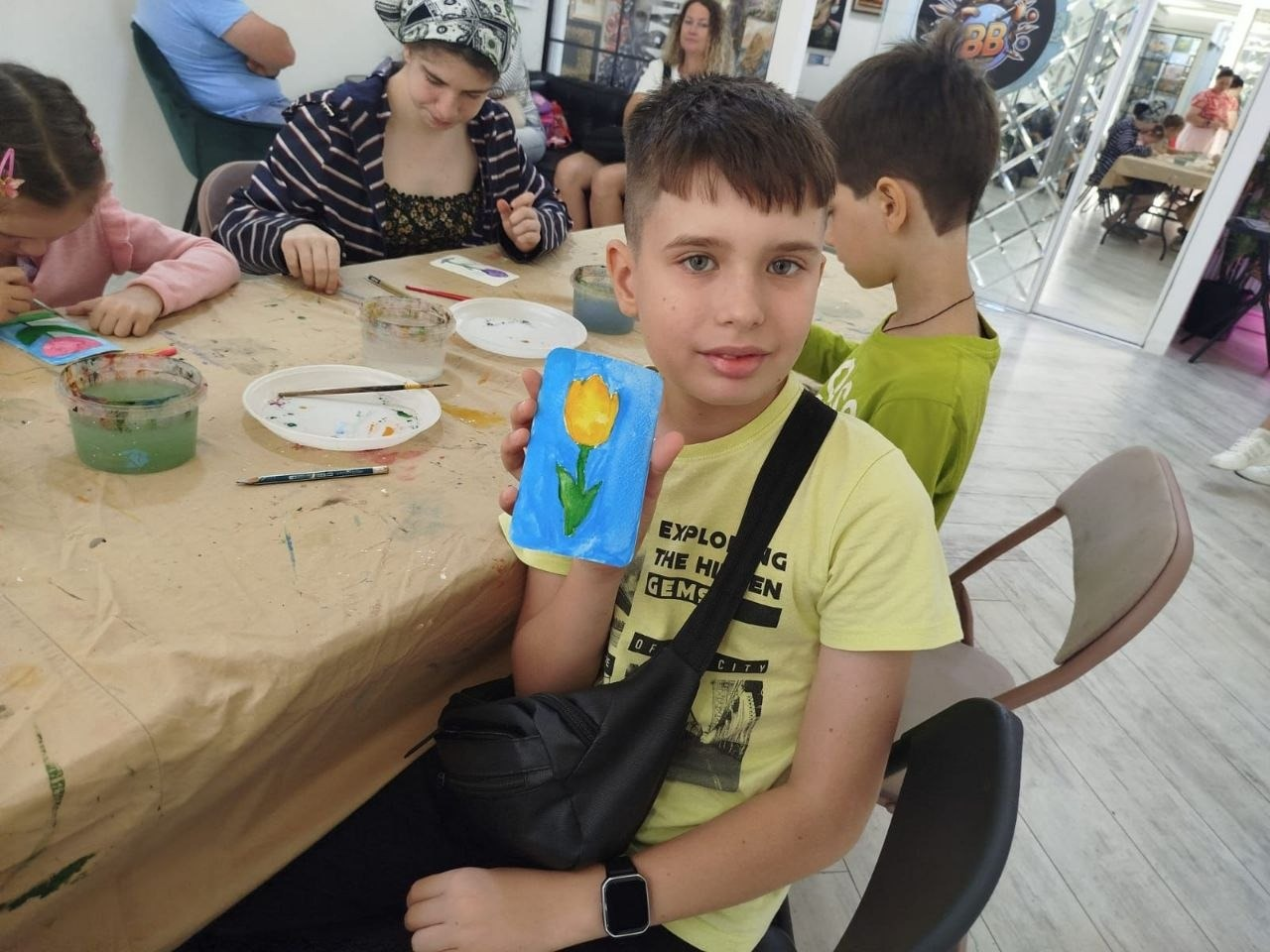 Благодійний художній майстер-клас для дітей-переселенців з Херсонщини в Одесі. На передньому плані крупним планом зображений хлопчик у світло-жовтій футболці з чорною сумкою-бананкою через плече, який сидить за столом і показує у руці створену ним листівку — намальований жовтий тюльпан на яскраво-блакитному тлі. Поруч за довгим столом, застеленим папером, інші діти також малюють листівки фарбами. На столі розкладені баночки з водою, пензлі та пластикові тарілки-палітри з розведеними фарбами. Ці творчі роботи будуть передані пацієнтам дитячої лікарні «Охматдит» для психологічної підтримки.