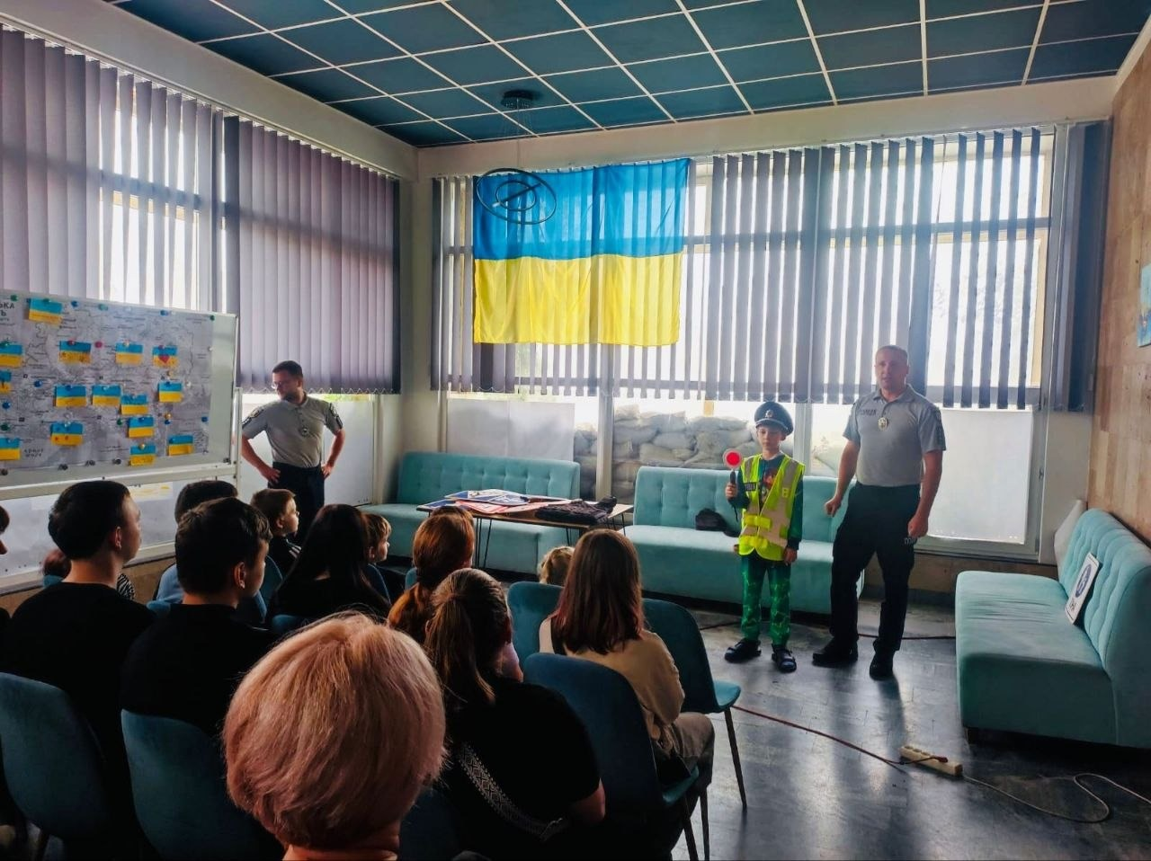 Зустріч дітей-переселенців із Херсонщини з патрульними поліцейськими у миколаївському координаційному центру «Вільні разом». У центрі зали біля великого вікна, де висить державний прапор України, стоїть хлопчик у світловідбивному жилеті та кашкеті патрульного, тримаючи в руці сигнальний диск регулювальника. Праворуч від нього стоїть поліцейський, який з посмішкою коментує процес екіпірування дитини. Ліворуч біля маркерної дошки з картою та синьо-жовтими наліпками стоїть інший патрульний, заклавши руки за спину. На передньому плані на стільцях сидить група дітей та дорослих, які спостерігають за демонстрацією.