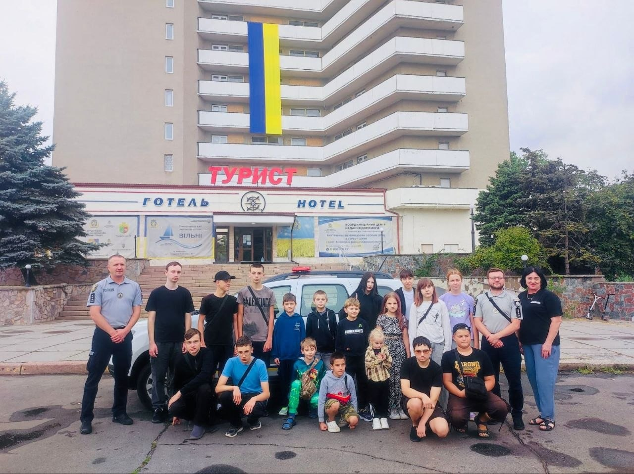 Спільне фото учасників зустрічі у миколаївському координаційному центрі «Вільні разом». Велика група дітей-переселенців із Херсонщини, їхніх батьків та патрульних поліцейських стоїть просто неба перед будівлею готелю «Турист». На передньому плані по центру кілька хлопчиків присіли навпочіпки, а дорослі та підлітки вишикувалися в ряд позаду них на тлі службового автомобіля поліції з проблисковими маячками. Крайній ліворуч стоїть патрульний у формі. На фасаді багатоповерхової будівлі готелю закріплено великий прапор України, а над входом розміщено банери координаційного центру.