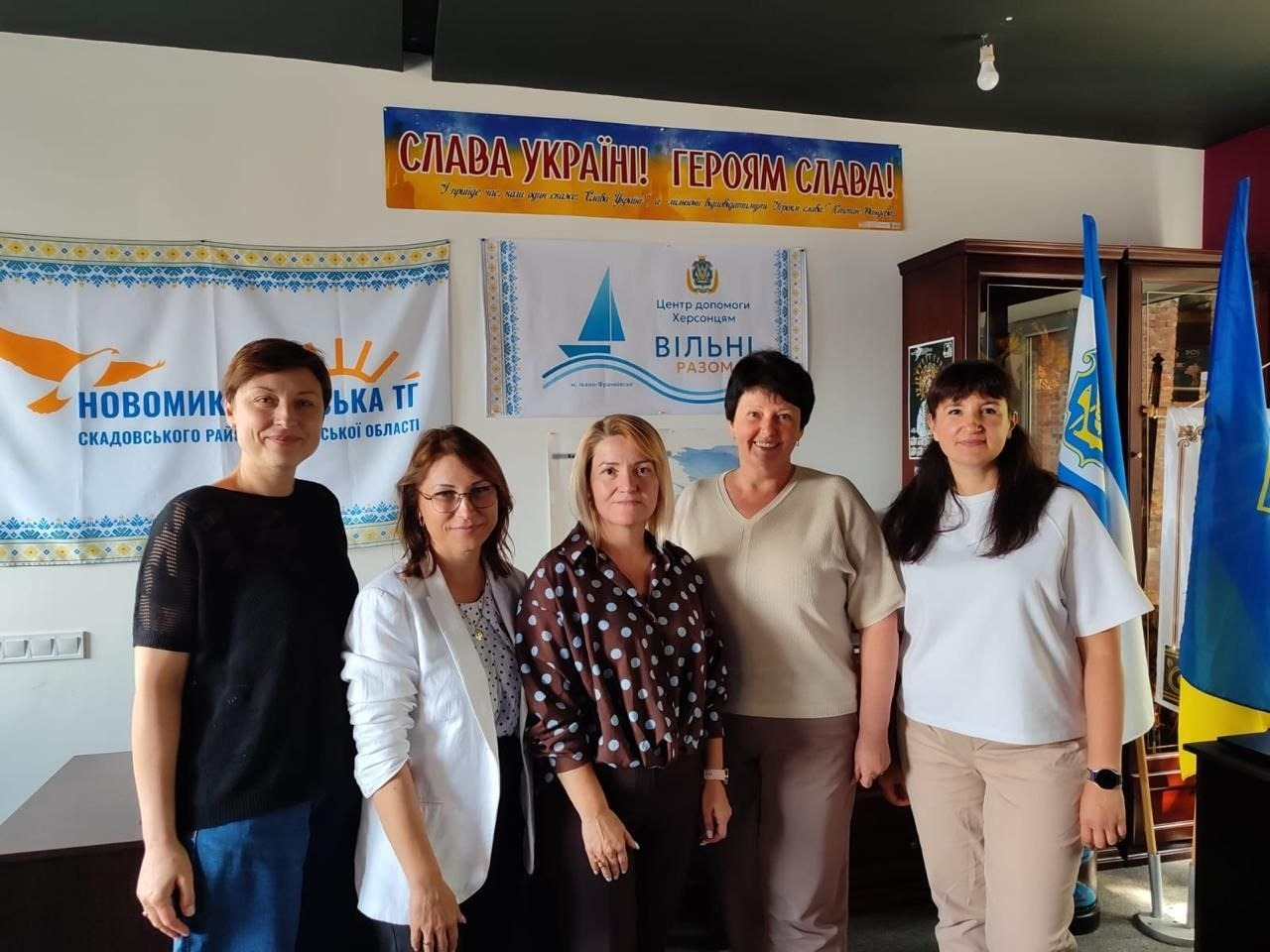 Робоча зустріч у координаційному центрі допомоги херсонцям «Вільні разом» в Івано-Франківську, присвячена підготовці статуту Новомиколаївської громади. У приміщенні центру стоять у ряд п'ять жінок — учасниць зустрічі, серед яких спеціалісти виконавчого комітету громади та фахівці Херсонського регіонального офісу GIZ. На задньому плані розвішані інформаційні банери, зокрема з написом «Новомиколаївська ТГ Скадовського району Херсонської області» та логотипом хабу «Вільні разом», а також закріплене патріотичне гасло «СЛАВА УКРАЇНІ! ГЕРОЯМ СЛАВА!». Праворуч на підставках встановлені державний прапор України та символіка громади.