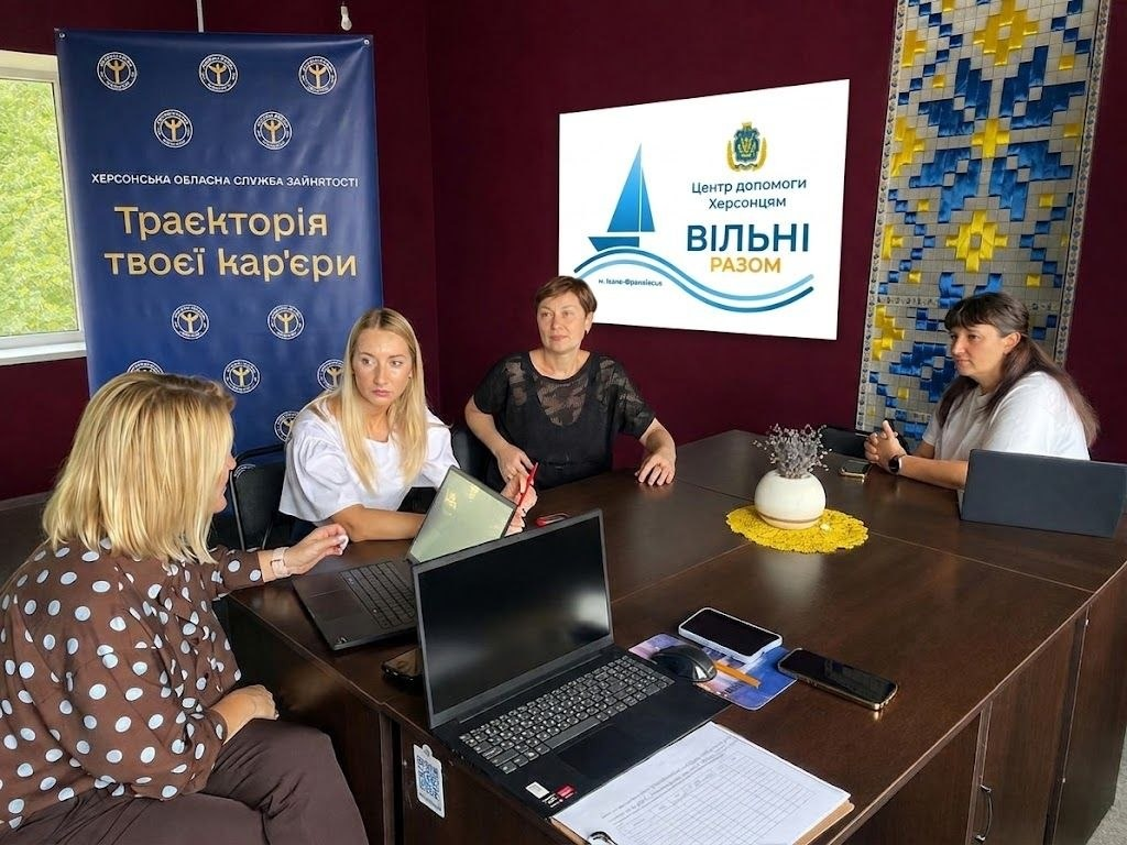 Робоча зустріч у координаційному центрі допомоги херсонцям «Вільні разом» в Івано-Франківську за участю представників Херсонської обласної служби зайнятості. За великим дерев'яним кутовим столом сидять чотири жінки. На столі перед ними відкриті ноутбуки, документи та смартфони. На задньому плані ліворуч розміщено синій банер Херсонської обласної служби зайнятості з написом «Траєкторія твоєї кар'єри». По центру розташовано білий екран із логотипом «Центр допомоги херсонцям ВІЛЬНІ РАЗОМ», а праворуч на стіні висить декоративне панно з традиційним українським орнаментом у синьо-жовтих кольорах. Учасниці зустрічі обговорюють робочі питання та взаємодію.