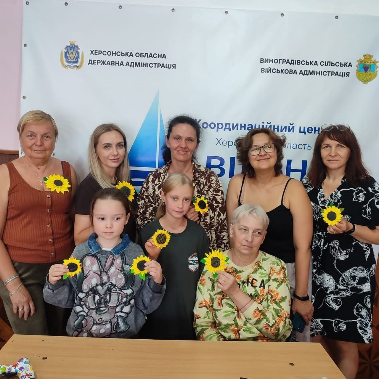 Майстер-клас «Виготовлення соняшника з фетру» до Дня пам'яті Захисників і Захисниць України, організований для внутрішньо переміщених осіб (ВПО) з Херсонщини у чернівецькому координаційному центрі «Вільні разом». Група з п'яти жінок різного віку та двох дівчаток стоїть біля столу в приміщенні хабу. Учасниці посміхаються в камеру та тримають у руках виготовлені власноруч квіти соняшника з яскраво-жовтими пелюстками — символи пам'яті за загиблими воїнами. На задньому плані видно великий білий банер із логотипом хабу та офіційними написами «Херсонська обласна державна адміністрація» та «Виноградівська сільська військова адміністрація».