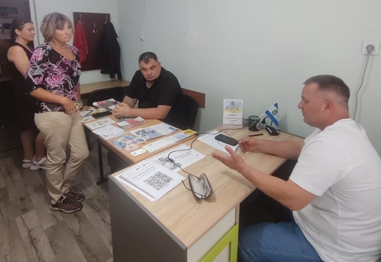 Особистий прийом внутрішньо переміщених осіб (ВПО) з Херсонщини, який провів начальник Новотроїцької селищної військової адміністрації Сергій Перетятько у чернівецькому координаційному центрі «Вільні разом». По центру за робочим столом сидить Сергій Перетятько у чорній футболці-поло, уважно вислуховуючи відвідувачів. Праворуч від нього за іншим столом сидить колега у білій футболці, який активно жестикулює під час надання роз'яснень. На столах розкладені різноманітні інформаційні буклети, брошури та пам'ятки з QR-кодами. Навпроти керівника стоять дві жінки-відвідувачки, які звернулися по допомогу та консультацію.