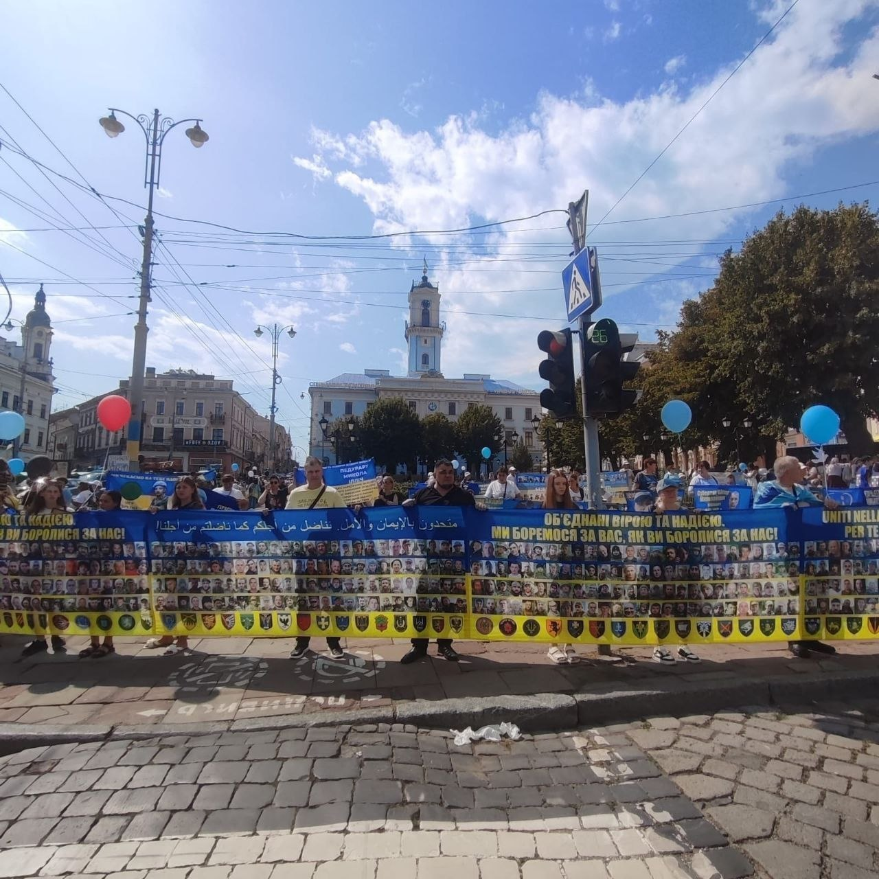 Участь фахівців чернівецького координаційного центру «Вільні разом» та внутрішньо переміщених осіб (ВПО) з Херсонщини в масштабній мирній акції на підтримку військовополонених та зниклих безвісти захисників. На Центральній площа Чернівців, на тлі міської Ратуші, учасники заходу тримають наддовгий банер із сотнями фотографій полонених і зниклих воїнів та емблемами бригад ЗСУ. На синій верхній частині банера великими жовтими літерами написані патріотичні гасла українською, арабською та іншими мовами, зокрема текст: «ОБ'ЄДНАНІ ВІРОЮ ТА НАДІЄЮ. МИ БОРЕМОСЯ ЗА ВАС, ЯК ВИ БОРОЛИСЯ ЗА НАС!». Люди стоять уздовж бруківки, тримаючи в руках сині та рожеві повітряні кульки під ясним небом із хмарами.