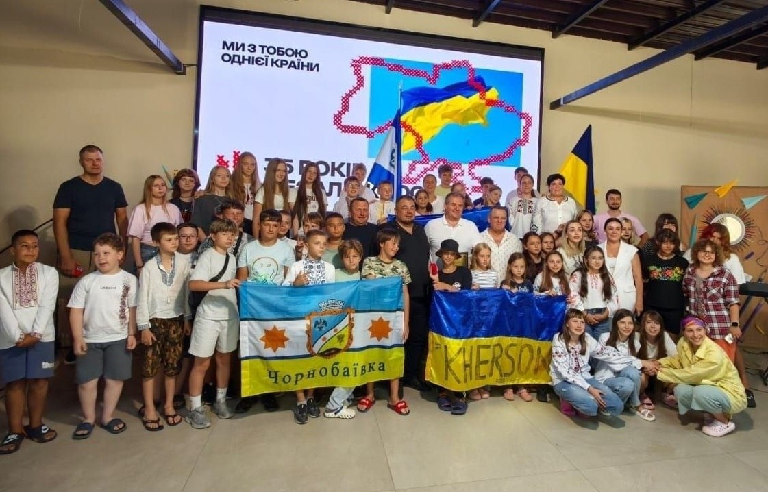 Спільне фото учасників патріотичного заходу в таборі «Your camp», організованого чернівецьким координаційним центром «Вільні разом» спільно з Палацом молоді і студентів Херсонської обласної ради. Велика група дітей-переселенців із Херсонщини різного віку, підлітків та педагогів вишикувалася разом для знімка. У самому центрі кадру серед дорослих стоїть начальник Новотроїцької СВА Сергій Перетятько у чорній футболці. Попереду учасники тримають два великі прапори: ліворуч — прапор Чорнобаївки з гербом громади та написом «Чорнобаївка», праворуч — синьо-жовтий прапор України з великим написом «KHERSON». Багато дітей одягнені у вишиванки. На задньому плані встановлено великий мультимедійний екран із картою України у патріотичних кольорах та налисом «МИ З ТОБОЮ ОДНІЄЇ КРАЇНИ» до Дня Незалежності.