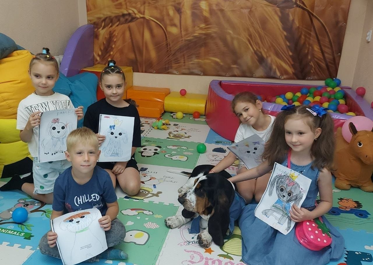 Заняття з психологічної допомоги «Курс емоційної стійкості» та «Будинок емоційної стійкості» для дітей-переселенців із Херсонщини в спортивно-оздоровчому таборі «Your Camp» у селі Ржавинці. На кольоровому м'якому килимку в ігровій кімнаті сидять п'ятеро усміхнених дітей, які тримають у руках свої малюнки з портретами собак. У центрі кадру разом із ними сидить велика чорно-біла собака породи бассет-гаунд у синій патріотичній бандані з тризубом, яка допомагає дітям у процесі психологічного розвантаження та каністерапії. На задньому плані видно сухий басейн із кольоровими кульками, м'які модулі та настінне панно. Захід організовано чернівецьким координаційним центром «Вільні разом».