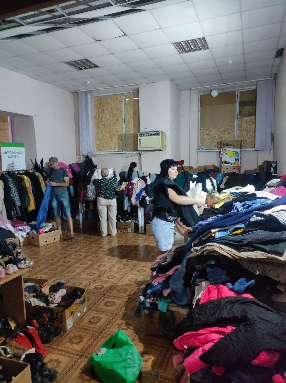 Переселенці з Херсонщини обирають речі у банку одягу, що діє в рамках соціальних сервісів координаційного центру «Вільні разом» у Миколаєві. У великому приміщенні на столах, вішалках-рейлах та в картонних коробках розкладено велику кількість вживаного одягу та взуття, якими скористалися 258 осіб. Кілька відвідувачів перебирають та розглядають речі. Вікна на задньому плані забиті захисними плитами ОСБ, на стіні видно кондиціонер, а ліворуч стоїть мобільний інформаційний стенд.
