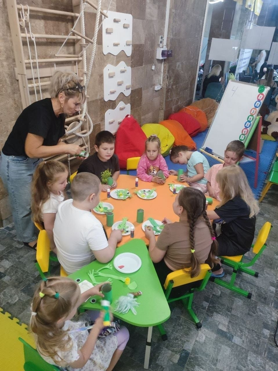 Діти-переселенці з Херсонщини беруть участь у патріотичному майстер-класі в координаційному центрі «Вільні разом» у Миколаєві, організованому до Дня Державного прапора та Дня Незалежності України [2026 рік]. Хлопчики та дівчатка сидять за яскравими кольоровими столами і створюють тематичні поробки із зеленого паперу та картону, використовуючи клей-олівці. Поруч стоїть вихователька, яка допомагає дітям у творчому процесі. На задньому плані дитячої кімнати видно шведську стінку, різнокольорові крісла-мішки та маркерну дошку з цифрами.