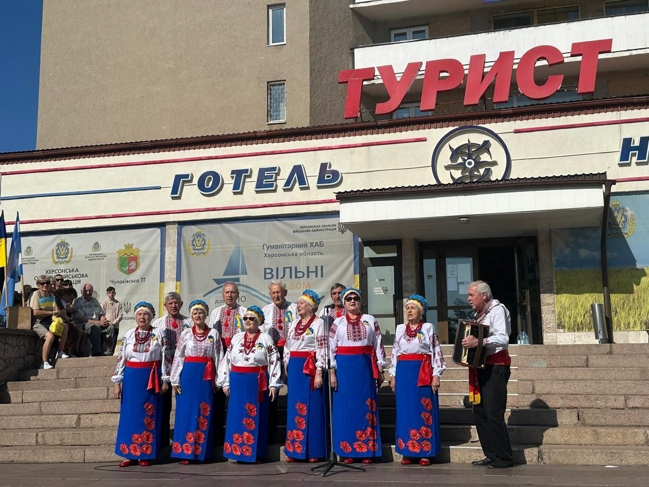 Український народний хор виступає на сходах перед будівлею готелю «Турист» у Миколаєві під час великого святкового заходу для ВПО, організованого координаційним центром «Вільні разом» до 35-ї річниці Незалежності України. Учасники ансамблю в патріотичних строях — білих вишиванках, синіх спідницях із квітковим візерунком та вінках — співають перед мікрофонами, а чоловік праворуч акомпанує їм на баяні. На фасаді будівлі над входом розміщено написи «ТУРИСТ» та «ГОТЕЛЬ». На стінах за артистами закріплені великі інформаційні банери Гуманітарного хабу Херсонської області «Вільні разом», Херсонської ОВА та Чулаківської територіальної громади. Ліворуч на сходах за концертом спостерігають глядачі.