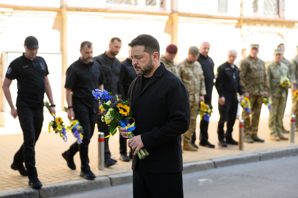 Президент України Володимир Зеленський тримає букет із соняшників та синьо-жовтих квітів під час хвилини мовчання у День пам’яті захисників України. Глава держави одягнений у чорне вбрання; його голова схилена, а очі заплющені в знак скорботи та вшанування подвигу полеглих воїнів біля Стіни пам’яті в Києві. На задньому плані вздовж тротуару вишикувалося вище військово-політичне керівництво країни, серед яких міністр оборони Євгеній Хмара, міністр внутрішніх справ Іван Вигівський, начальник Генштабу ЗСУ Ігор Скибюк, керівник Офісу Президента Кирило Буданов, секретар РНБО Ігор Клименко, голова СЗР Рустем Умєров та керівник ГУР Олег Іващенко, а також військовослужбовці в камуфляжній формі. Подія відбувається під сонячним світлом на тлі світлої стіни будівлі.