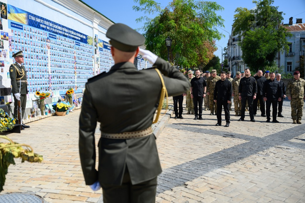 Президент України Володимир Зеленський разом із вищим військово-політичним керівництвом країни вшановує хвилиною мовчання подвиг полеглих героїв у День пам’яті захисників України. Ліворуч на Михайлівській площі розташована Стіна пам’яті полеглих захисників і захисниць України в російсько-українській війні, заповнена численними фотографіями; біля її підніжжя встановлено кошики з квітами, соняшники та лампадки. Поруч у парадній формі стоїть військовослужбовець почесної варти, а на першому плані спиною до камери інший військовий віддає честь. Глава держави та посадовці, серед яких міністр оборони Євгеній Хмара, міністр внутрішніх справ Іван Вигівський, начальник Генштабу ЗСУ Ігор Скибюк, керівник Офісу Президента Кирило Буданов, секретар РНБО Ігор Клименко, голова СЗР Рустем Умєров та керівник ГУР Олег Іващенко, вишикувалися на бруківці на тлі зелених дерев.