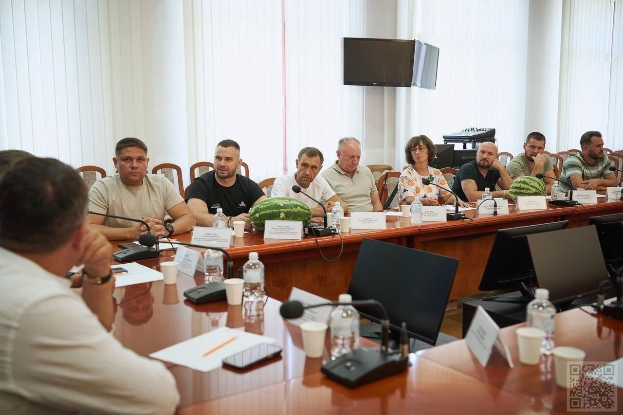 Робоча зустріч у залі засідань за великим дерев'яним столом. На першому плані ліворуч спиною до камери сидить чоловік у білій сорочці. Навпроти нього за столом сидить ряд учасників зустрічі. Серед них (зліва направо): директор Департаменту розвитку сільського господарства та зрошення Херсонської ОВА Дмитро Юнусов у світлій бежевій футболці; голова асоціації фермерів та приватних землевласників Херсонської області Віктор Гордієнко в чорній футболці; чоловік у білій футболці та інші представники аграрного сектору. На столі перед ними встановлено іменні таблички, конференц-мікрофони, розкладено папери, паперові стаканчики та пляшки з водою. Також на столі лежать два великі стиглі кавуни. На задньому плані видно великі вікна з вертикальними білими жалюзі, вимкнений телевізор на стіні та музичне обладнання у кутку кімнати. У правому нижньому кутку зображення розміщено невеликий QR-код.