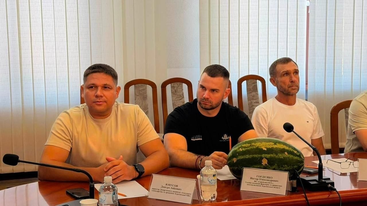 Представники аграрного сектору Херсонщини під час зустрічі в Рівненській ОВА в межах робочого візиту для обговорення міжрегіональної співпраці та підтримки бізнесу. За дерев'яним столом на тлі вікна з білими вертикальними жалюзі сидять троє чоловіків. Ліворуч — директор Департаменту розвитку сільського господарства та зрошення Херсонської ОВА Дмитро Юнусов у світло-бежевій футболці; перед ним стоїть іменна табличка. Посередині сидить голова асоціації фермерів та приватних землевласників Херсонської області Віктор Гордієнко в чорній футболці, який уважно слухає і дивиться ліворуч. Праворуч від нього сидить представник агросектору у білій футболці. На столі перед ними лежить великий стиглий кавун — символічний подарунок від херсонської делегації, біля якого встановлено відповідну табличку з іменем Віктора Гордієнка. Також на столі розміщено конференц-мікрофони та пляшку води.