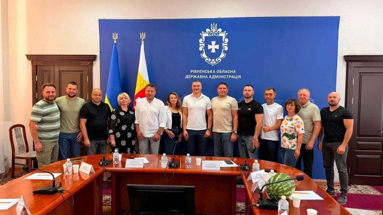 У залі засідань Рівненської ОДА учасники зустрічі стоять в один ряд для спільного фото перед великим синім банером установи. У центрі ряду стоїть заступник голови Рівненської ОДА Олександр Кохан у білій футболці-поло. Ліворуч від нього — народний депутат України Дмитро Соломчук у білій сорочці, праворуч — директор Департаменту агророзвитку Херсонської ОВА Дмитро Юнусов у світло-бежевій футболці та голова асоціації фермерів Херсонщини Віктор Гордієнко в чорній футболці. По обидва боки від них розмістилися інші представники агросектору Херсонщини та БФ «Жнива Перемоги». Ліворуч стоять державний прапор України та прапор Рівненської області. На першому плані на столі засідань лежить великий смугастий херсонський кавун.