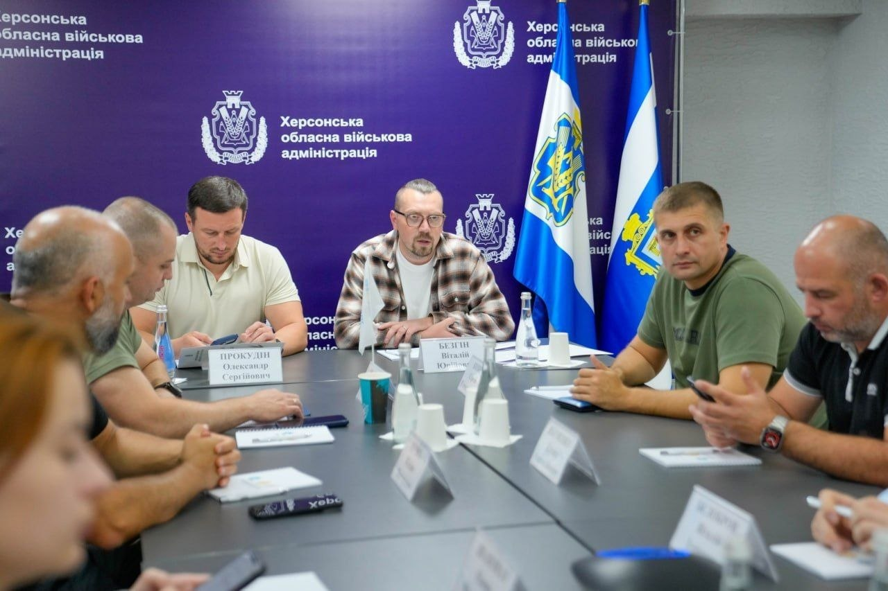 Робоча нарада на чолі з Міністром у справах громад, територій та внутрішньо переміщених осіб Віталієм Безгіном та начальником Херсонської ОВА Олександром Прокудіним у залі засідань ХОВА. Засідання присвячене розробці драфту законопроєкту про соціальні гарантії для працівників прифронтових територій та забезпеченню ВПО тимчасовим житлом і виплатами. Учасники зустрічі сидять навпроти один одного за довгим сірим столом. З лівого боку столу по центру в окулярах та сорочці-кардигані у коричневу клітинку сидить очільник міністерства Віталій Безгін. Поруч із ним ліворуч у світлій футболці-поло розмістився начальник Херсонської ОВА Олександр Прокудін. Далі ліворуч від Прокудіна сидить начальник Херсонської міської військової адміністрації Ярослав Шанько, а ще лівіше, на самому передньому плані ближче до камери — заступник голови Херсонської РДА Юліан Лучко. Перед посадовцями встановлено іменні таблички. З правого боку столу навпроти них сидить народний депутат України Сергій Козир в оливково-зеленій футболці та інші керівники військових адміністрацій населених пунктів області. На задньому плані розташовано великий фіолетовий банер із написом Херсонська обласна військова адміністрація, поруч із яким стоять державний прапор України та офіційний прапор Херсонської області. На столі розкладено робочі документи, ручки, стоять пляшки з водою та паперові стаканчики.