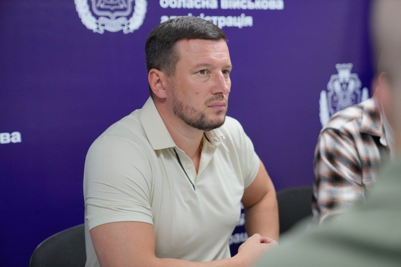 Портретний знімок начальника Херсонської обласної військової адміністрації Олександра Прокудіна під час спільної робочої наради з Міністром у справах громад, територій та внутрішньо переміщених осіб Віталієм Безгіном. Олександр Прокудін сидить за столом у світлій футболці-поло та уважно слухає виступ колег, присвячений розробці законопроєкту про соціальні гарантії для працівників прифронтових територій та житловій підтримці переселенців. На задньому плані розташовано фіолетовий бренд-волл із написами обласна військова адміністрація та гербом установи. Праворуч на задньому плані видно частину плеча міністра Віталія Безгіна в сорочці в коричневу клітинку, а на передньому плані — розмитий силует іншого учасника засідання.