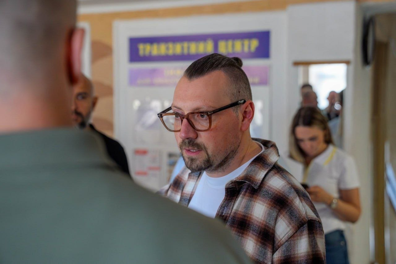 Міністр у справах громад, територій та внутрішньо переміщених осіб Віталій Безгін відвідує транзитний центр на Херсонщині для людей, які евакуюються з небезпечних територій. Міністр зображений крупним планом в окулярах та сорочці в коричневу клітинку поверх білої футболки під час спілкування з начальником Херсонської ОВА Олександром Прокудіним, якого видно зі спини на першому плані ліворуч. На задньому плані у розмитому фокусі розташовано інформаційну вивіску з написом Транзитний центр, а також видно інших співробітників установи та супроводжуючих осіб у світлому приміщенні хабу.