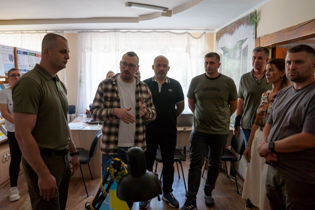 Робоча група посадовців обговорює виклики та умови перебування евакуйованих громадян у приміщенні місця тимчасового проживання на Херсонщині. Ліворуч на першому плані стоїть начальник Херсонської міської військової адміністрації Ярослав Шанько в оливковій футболці-поло. Поруч із ним по центру активно жестикулює під час розмови Міністр у справах громад, територій та внутрішньо переміщених осіб Віталій Безгін в окулярах та сорочці в коричневу клітинку. Навпроти них із правого боку стоять народний депутат України Сергій Козир у темно-зеленій футболці Ami Paris та перший заступник начальника Херсонської міської військової адміністрації Михайло Линецький у сіро-зеленій футболці на передньому плані праворуч. Позаду посадовців у кімнаті перебувають інші люди, серед яких чоловік у чорній сорочці-поло, жінка у світлій блузі та колеги з координаційного хабу. На тлі кімнати видно велике вікно з білою завісою, робочі столи та інформаційні стенди для ВПО на стіні ліворуч.