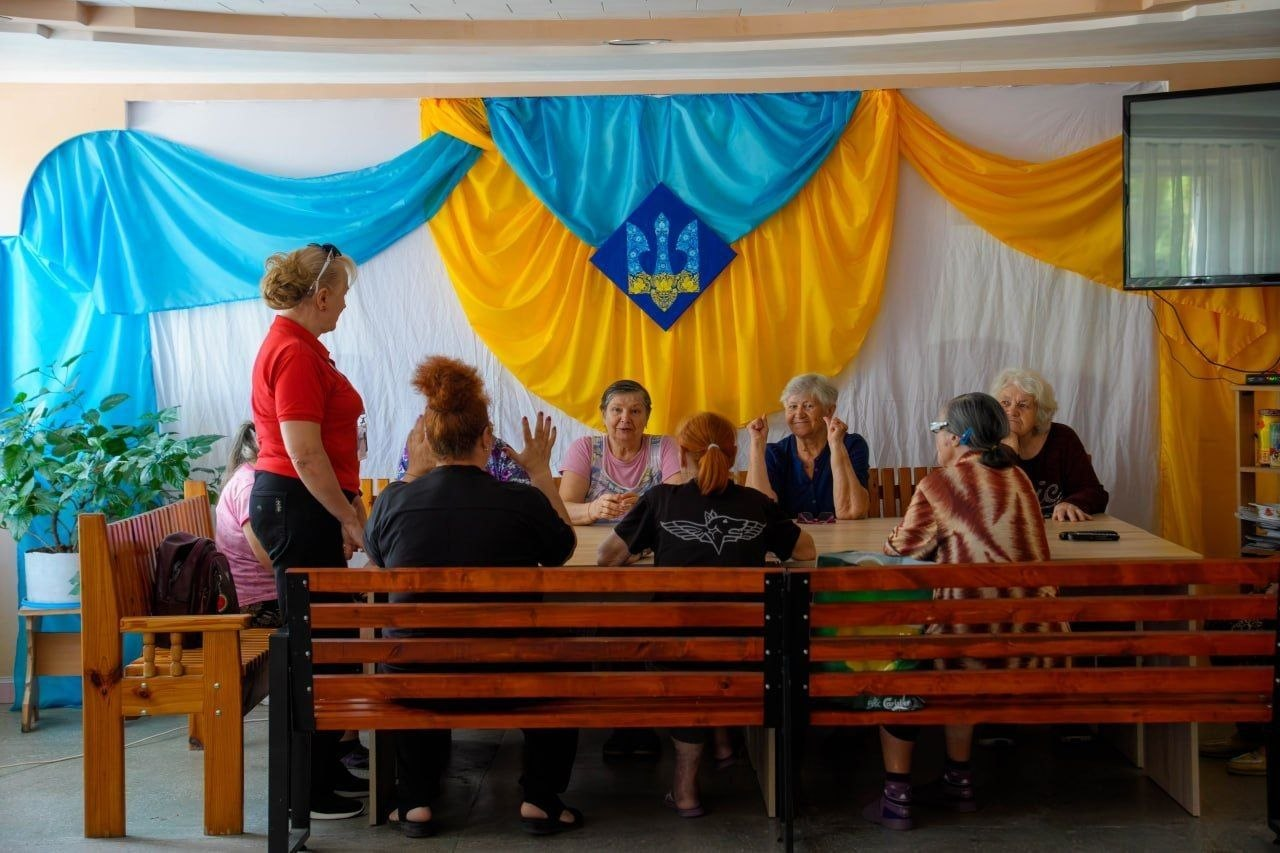 Внутрішньо переміщені особи та евакуйовані жителі спілкуються під час візиту робочої групи у місці тимчасового проживання на Херсонщині. За довгим дерев'яним столом сидить група літніх жінок із числа ВПО, які жваво обговорюють свої потреби та запити. Ліворуч біля столу стоїть представниця установи в червоній футболці, яка координує діалог. На задньому плані вся стіна прикрашена великим синьо-жовтим патріотичним драпуванням із тканини, у центрі якого закріплено малий державний герб України. Ліворуч у кутку кімнати стоїть пишний зелений кімнатний вазон, а праворуч під стелею встановлено телевізійний екран. Перед столом розташовані дерев'яні лавки зі спинками.