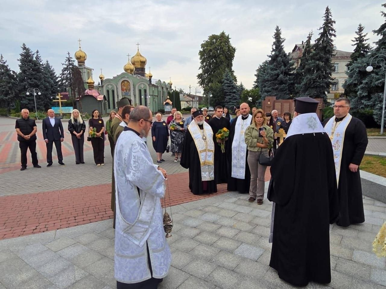 На фотографії зафіксовано молебень у пам’ять полеглих Захисників та Захисниць України, який відвідали представники Чаплинської, Долматівської, Білолуцької, Попаснянської та Черкаської громад за підтримки координаційного центру «Вільні разом» у Черкасах. На брукованій площі перед храмом із золотими куполами священнослужителі у святкових ризах проводять релігійну церемонію, де один із кліриків тримає кадило, а ієрарх у чорній мантії та білому куколі стоїть спиною до камери. Навколо духовенства на тлі високих ялин зібралися представники громад та парафіяни в цивільному одязі, які молитовно вшановують пам'ять загиблих героїв України.