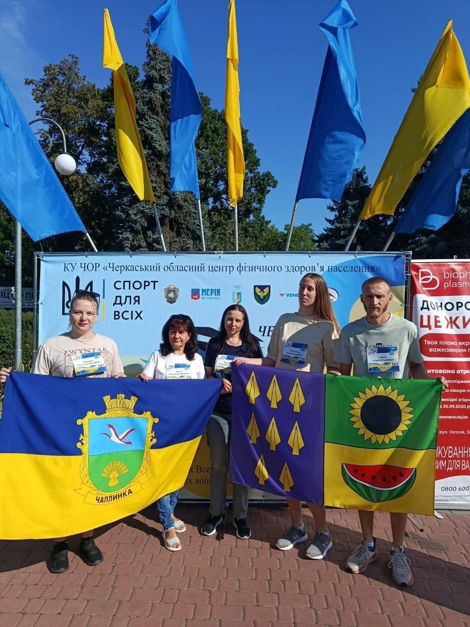 На фотографії зафіксовано групу херсонських переселенців з координаційного центру «Вільні разом» у Черкасах, які взяли участь у щорічному всеукраїнському забігу «Шаную воїнів – біжу за героїв України». П'ятеро учасників у спортивному одязі та з нагрудними номерами забігу стоять просто неба на тлі банера «Черкаського обласного центру фізичного здоров’я населення» під майоріючими синьо-жовтими прапорами України. Вони гордо розгортають офіційні прапори своїх рідних громад Херсонщини: ліворуч дві жінки тримають синьо-жовтий прапор селища Чаплинка з його гербом, по центру дівчата розгортають темно-синій прапор Долматівської громади із золотими наконечниками стріл, а чоловік праворуч тримає зелено-жовтий прапор Бехтерської громади із зображенням соняшника та скибки кавуна.