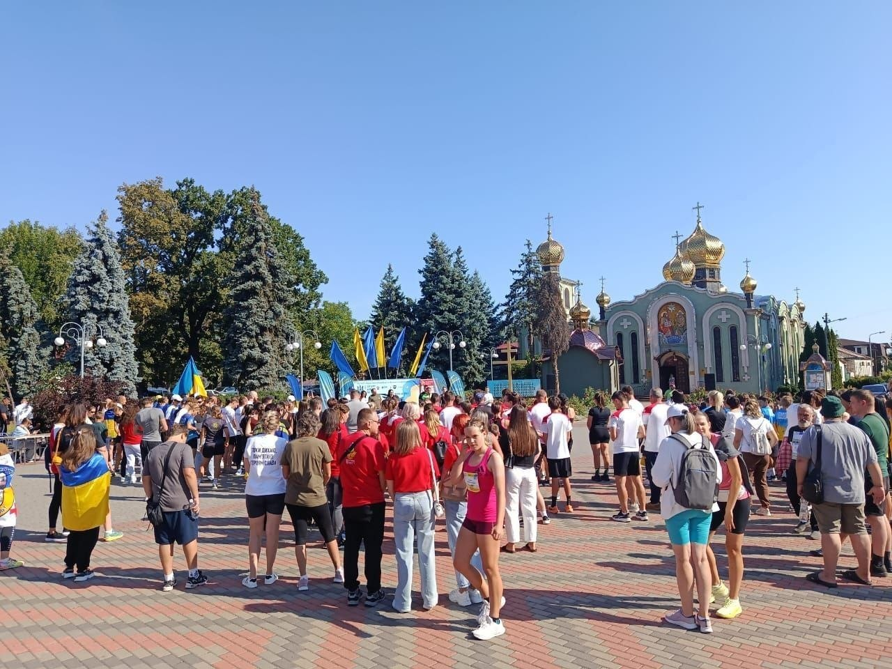 На фотографії зафіксовано участь херсонських переселенців з координаційного центру «Вільні разом» в урочистому патріотичному заході до Дня Незалежності України, організованому Черкаською обласною та міською радами. Велика кількість учасників у спортивному та повсякденному одязі з патріотичною символікою зібралася на великій брукованій площі перед храмом із золотими куполами під ясним синім небом. На задньому плані, біля зеленої зони з високими ялинами, майоріють державні синьо-жовті прапори України на флагштоках, де облаштовано офіційну зону зборів, а серед людей на площі видно розгорнутий український стяг.