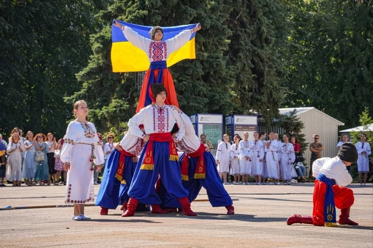 На фотографії зафіксовано хореографічний виступ танцювального колективу під час урочистого заходу до Дня Незалежності України, організованого Черкаською обласною та міською радами, який відвідали херсонські переселенці з координаційного центру «Вільні разом». У центрі майданчика просто неба артисти у вишиванках та синіх шароварах утворюють динамічну трирівневу фігуру, де на вершині козак у шапці тримає широко розгорнутий державний синьо-жовтий прапор України. Ліворуч на передньому плані стоїть дівчина в білій вишитій сукні, праворуч на коліні схилився юний танцюрист, а на задньому плані за патріотичним виступом на тлі зелених дерев спостерігають численні глядачі та учасники свята.