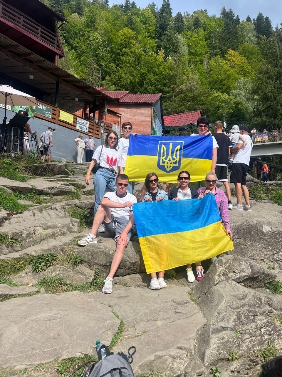 На фотографії зафіксовано групу херсонських переселенців із координаційного центру «Вільні разом» в Івано-Франківську під час культурно-просвітницької поїздки до міста Яремче, організованої з нагоди Дня Державного прапора України та 35-ї річниці Незалежності України. Семеро учасників подорожі у літньому повсякденному одязі та сонцезахисних окулярах позируют на скелястому березі біля водоспаду, гордо розгортаючи два державні синьо-жовті стяги, один з яких прикрашений золотим тризубом по центру. На задньому плані кадру на тлі густого зеленого лісу та гірського схилу видно дерев'яні відпочинкові комплекси, тераси та інших туристів.