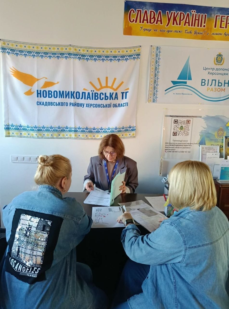 На фотографії зафіксовано процес надання правової допомоги внутрішньо переміщеним особам із Херсонщини в координаційному центрі «Вільні разом» в Івано-Франківську, де юрист благодійного фонду «Право на захист» надав 2 індивідуальні консультації. За робочим столом у світлій кімнаті сидить жінка-юрист в окулярах та сірому піджаку, яка уважно вивчає документи у світло-зеленій папці-швидкозшивачі. Навпроти неї спиною до камери сидять дві жінки-відвідувачки в джинсових куртках, які ознайомлюються з юридичними бланками. Консультація проходить на тлі стіни з патріотичним плакатом «СЛАВА УКРАЇНІ! ГЕРОЯМ СЛАВА!» та банерами Новомиколаївської ТГ Скадовського району й центру допомоги херсонцям.