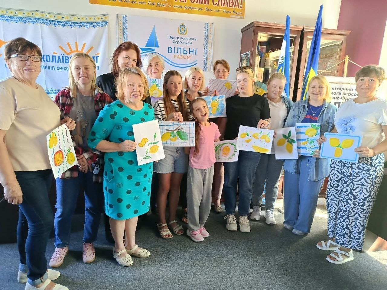 На фотографії зафіксовано велику групу херсонських переселенок (жінок та дітей), які зібралися разом у приміщенні координаційного центру «Вільні разом» в Івано-Франківську після завершення творчого майстер-класу з малювання та з усмішками демонструють свої готові художні роботи. Учасниці тримають у руках малюнки, на яких зображені яскраві жовті лимони та інші соковиті фрукти, виконані у різних художніх стилях. Вони стоять у світлій кімнаті на тлі державних прапорів України та офіційних банерів із символікою Новомиколаївської ТГ Скадовського району й гуманітарного центру допомоги херсонцям.
