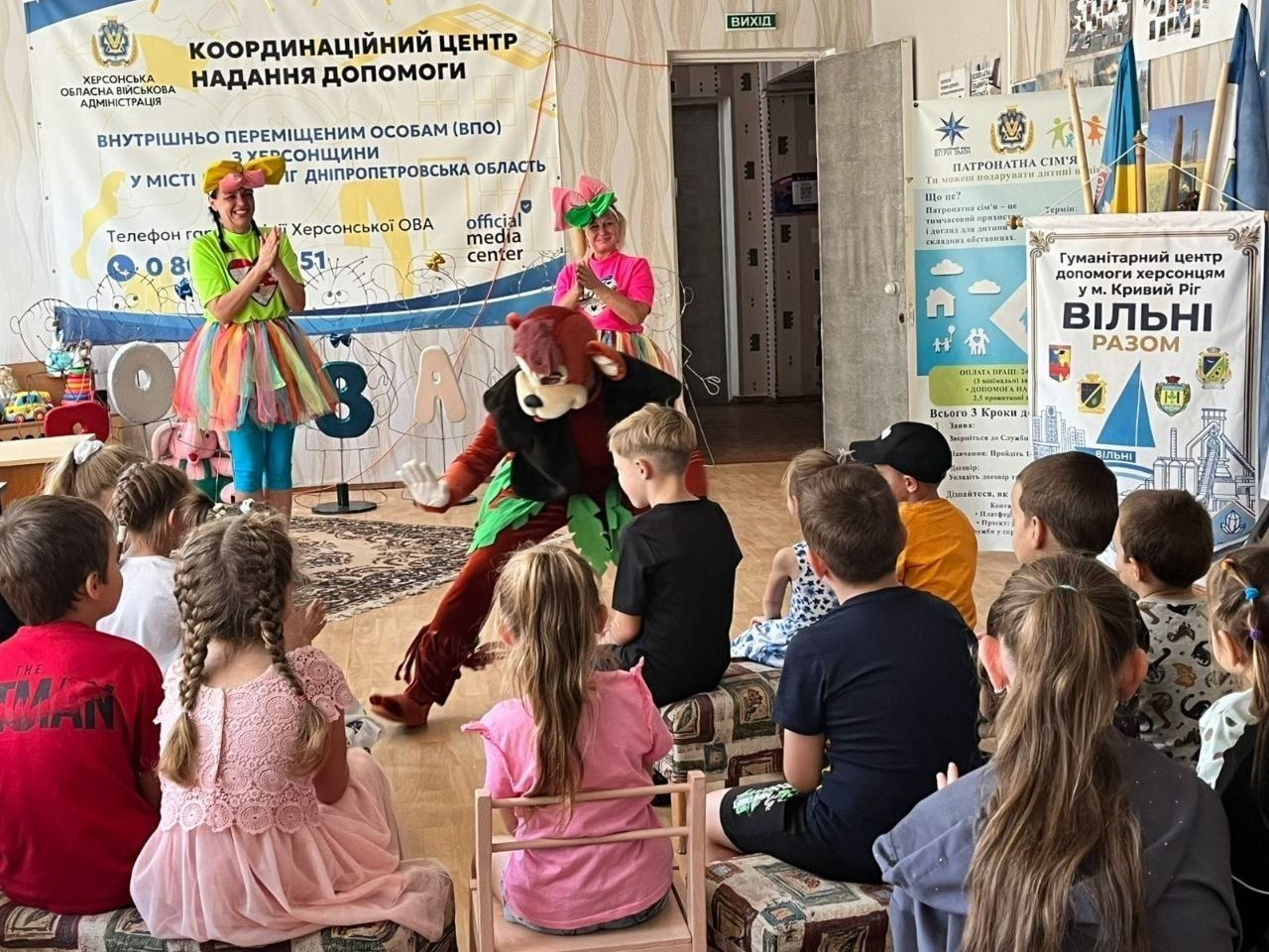 На фотографії зафіксовано інтерактивну виставу «GAME-сейшн» від акторів Криворізького міського театру ляльок, організовану для юних херсонських переселенців у координаційному центрі «Вільні разом» у Кривому Розі. Перед групою дітей, які сидять півколом спиною до камери, виступають троє артистів у яскравих сценічних образах: дві жінки-аніматорки в різнокольорових спідницях-пачках та актор у костюмі ведмедя, який присів на підлогу для гри з малечею. Подія проходить на тлі стіни з банерами Херсонської ОВА «Координаційний центр надання допомоги ВПО з Херсонщини у місті Кривий Ріг», інформаційними стендами «Гуманітарний центр допомоги херсонцям у м. Кривий Ріг ВІЛЬНІ РАЗОМ» та матеріалами про патронатні сім'ї.