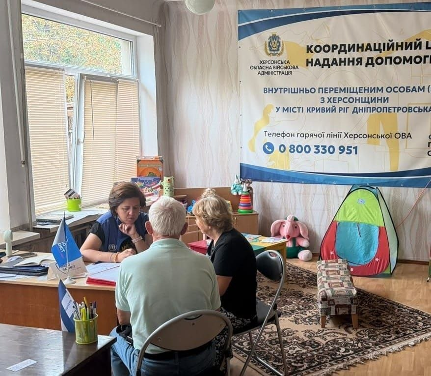 На фотографії зафіксовано процес надання комплексної допомоги внутрішньо переміщеним особам із Херсонщини від фахівців провідних гуманітарних організацій у координаційному центрі «Вільні разом» у Кривому Розі. У світлій кімнаті хабу за робочим столом сидіти представниця однієї з партнерських організацій у фірмовому темно-синьому жилеті, яка проводить індивідуальну консультацію для літньої пари переселенців. Зустріч, під час якої 15 людей отримали підтримку від БФ «Право на захист», ГО «Десяте квітня», БО «Схід SOS» та ГМ «Проліска», проходить біля вікна на тлі великого офіційного банера Херсонської ОВА «Координаційний центр надання допомоги ВПО з Херсонщини у місті Кривий Ріг» та дитячої ігрової зони з іграшками й кольоровим наметом.