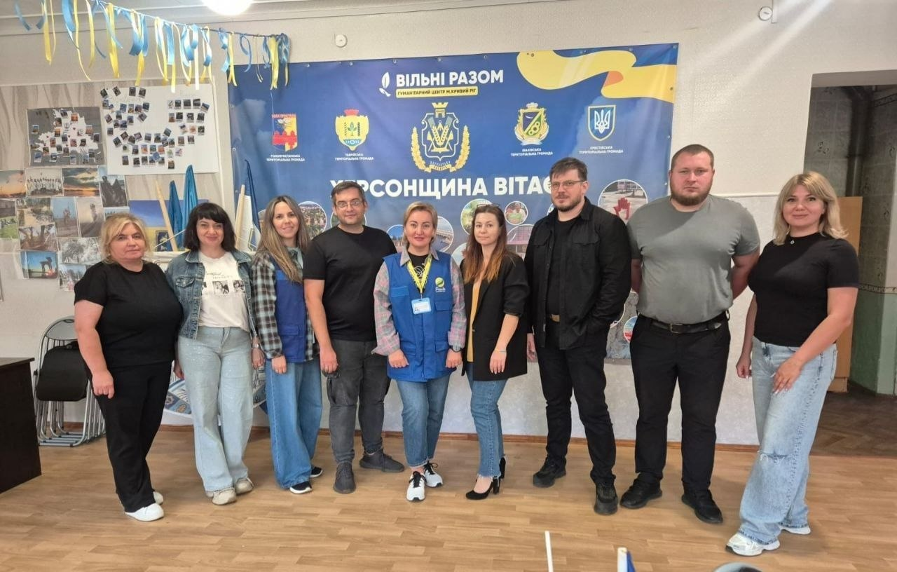 На фотографії зафіксовано колективне групове фото працівників та партнерів координаційного центру «Вільні разом» у Кривому Розі, який надає системну підтримку внутрішньо переміщеним особам із Херсонщини. Дев'ять представників гуманітарного хабу та партнерських організацій стоять в один ряд у світлій кімнаті, дві фахівчині по центру одягнені у фірмові сині жилети «Проліска» з логотипом УВКБ ООН. Вони позирують на тлі великого офіційного банера з емблемами деокупованих громад та написами «ХЕРСОНЩИНА ВІТАЄ» та «ВІЛЬНІ РАЗОМ. Гуманітарний центр допомоги херсонцям у м. Кривий Ріг», а ліворуч на стіні під патріотичними стрічками розміщено фотостенд із хронікою роботи хабу.