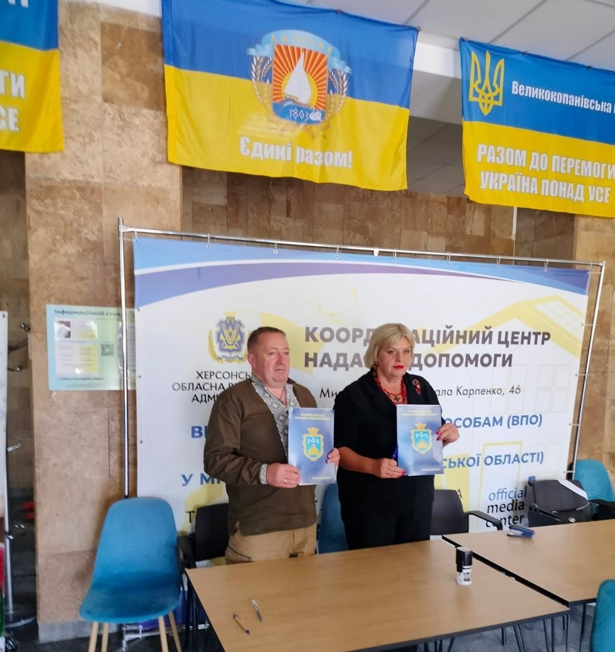 На фотографії зафіксовано підписання меморандуму про співпрацю між начальником Чулаківської СВА Скадовського району Херсонської області Русланом Русалем та головою Курісівської громади Березівського району Одеської області Наталією Бугайчук у месах проєкту «Пліч-о-пліч: згуртовані громади» на базі координаційного центру «Вільні разом» у Миколаєві. Чоловік та жінка тримають у руках офіційні сині папки з гербом Херсонщини, позируючи перед робочими столами на тлі великого банера Херсонської ОВА «Координаційний центр надання допомоги ВПО з Херсонської області». Над ними на кам'яній стіні розвішано великі синьо-жовті прапори України, зокрема прапор Великокопанівської громади Херсонської області з гаслом «Єдині разом!» та прапор із написом «Разом до перемоги! Україна понад усе».