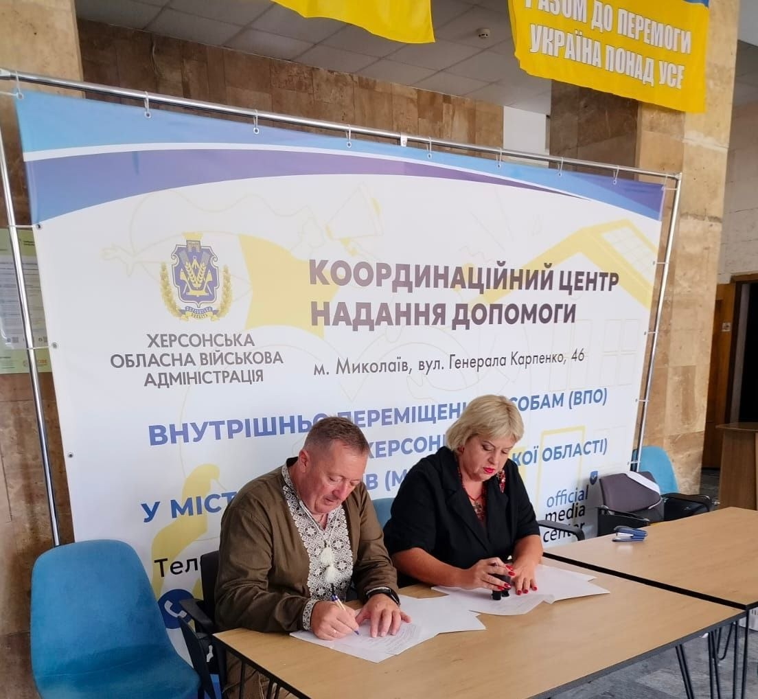 На фотографії зафіксовано процес безпосереднього підписання меморандуму про співпрацю між начальником Чулаківської СВА Скадовського району Херсонської області Русланом Русалем та головою Курісівської громади Березівського району Одеської області Наталією Бугайчук у межах проєкту «Пліч-о-пліч: згуртовані громади» на базі координаційного центру «Вільні разом» у Миколаєві. Чоловік у вишиванці та коричневому піджаку й жінка у чорній сукні зосереджено підписують офіційні документи, сидячи за дерев'яним столом на тлі великого банера Херсонської ОВА «Координаційний центр надання допомоги ВПО з Херсонської області». Над посадовцями на кам'яній стіні хабу розвішано великі синьо-жовті прапори України, зокрема прапор Великокопанівської громади Херсонської області з гаслом «Єдині разом!» та прапор із патріотичним написом «Разом до перемоги! Україна понад усе».