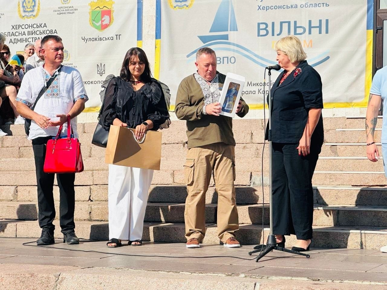 На фотографії зафіксовано урочисту частину заходу у Миколаєві з нагоди підписання меморандуму про співпрацю між Чулаківською СВА Скадовського району Херсонської області на чолі з Русланом Русалем та Курісівською громадою Березівського району Одеської області на чолі з Наталією Бугайчук у межах проєкту «Пліч-о-пліч: згуртовані громади». На відкритій сцені перед кам'яними сходами очільники громад та представники координаційного центру «Вільні разом» у патріотичному вбранні та вишиванках здійснюють обмін символічними подарунками. Голова Курісівської громади Наталія Бугайчук у чорному костюмі виступає перед мікрофоном, начальник Чулаківської СВА Руслан Русаль у зеленій вишиванці тримає святковий сувенір, а ліворуч стоять представники хабу з подарунковим крафтовим пакетом. Подія проходить під час відзначення 35-ї річниці Незалежності України на тлі великого світлого банера з синім вітрильником, написом «Херсонська область. ВІЛЬНІ РАЗОМ» та офіційними гербами Чулаківської й Великокопанівської територіальних громад.