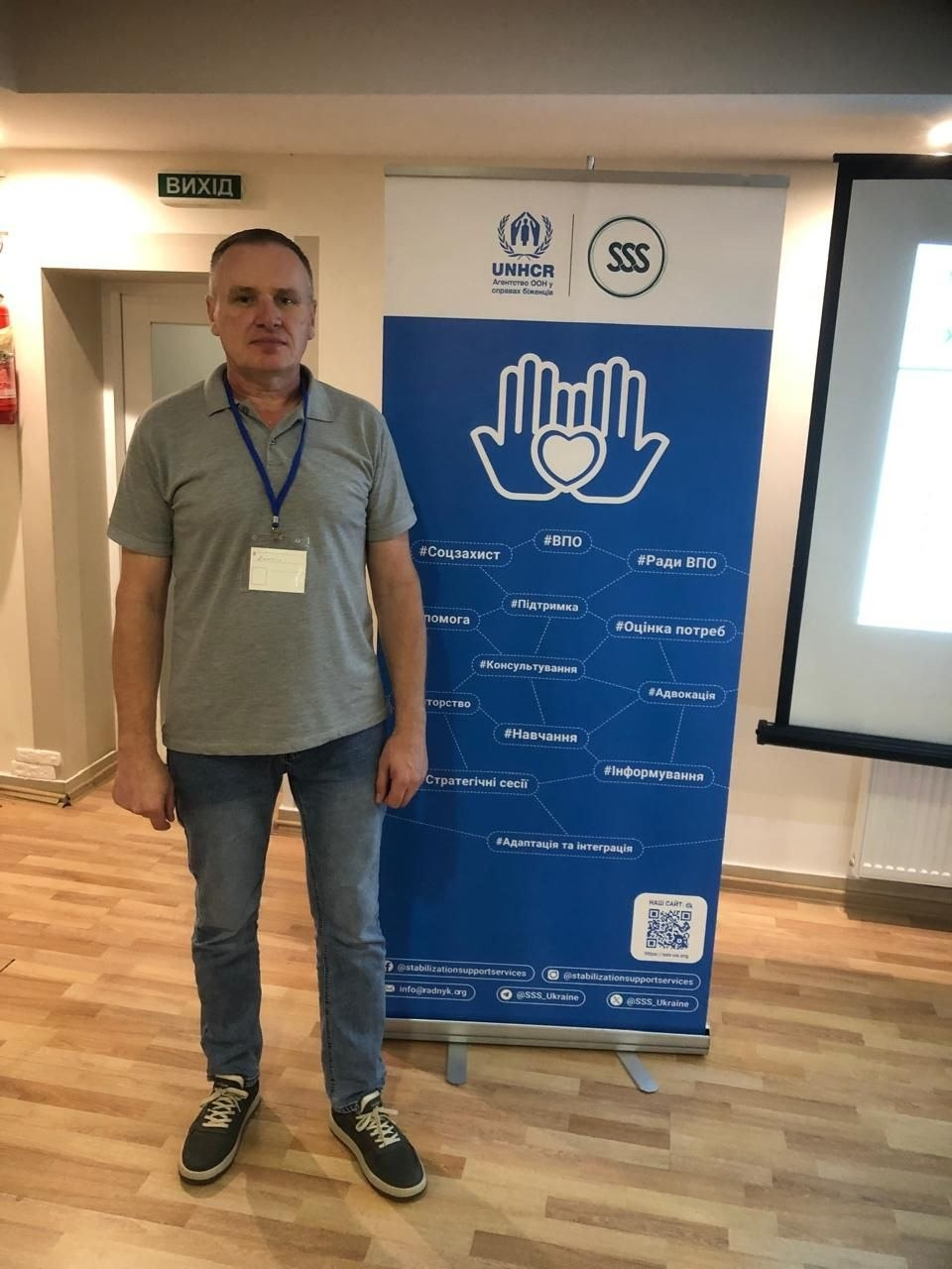 На фотографії зафіксовано начальника Рубанівської СВА Каховського району Херсонської області Дмитра Безрідного із бейджем на шиї, який у сірій футболці-поло, синіх джинсах та кросівках позирує під час зустрічі з обміну досвідом між радами ВПО у Житомирі. Він стоїть у приміщенні на тлі великого синього інформаційного стенда, організованого БО «БФ «Стабілізейшен Суппорт Сервісез» (SSS) за підтримки УВКБ ООН (UNHCR). На банері розміщено емблему у вигляді долонь із серцем, посилання на соцмережі та тематичні хештеги, серед яких «ВПО», «Ради ВПО», «Соцзахист» та «Консультування», а на задньому плані кімнати помітно білі двері з табличкою «ВИХІД» та частину великого проекційного екрана.