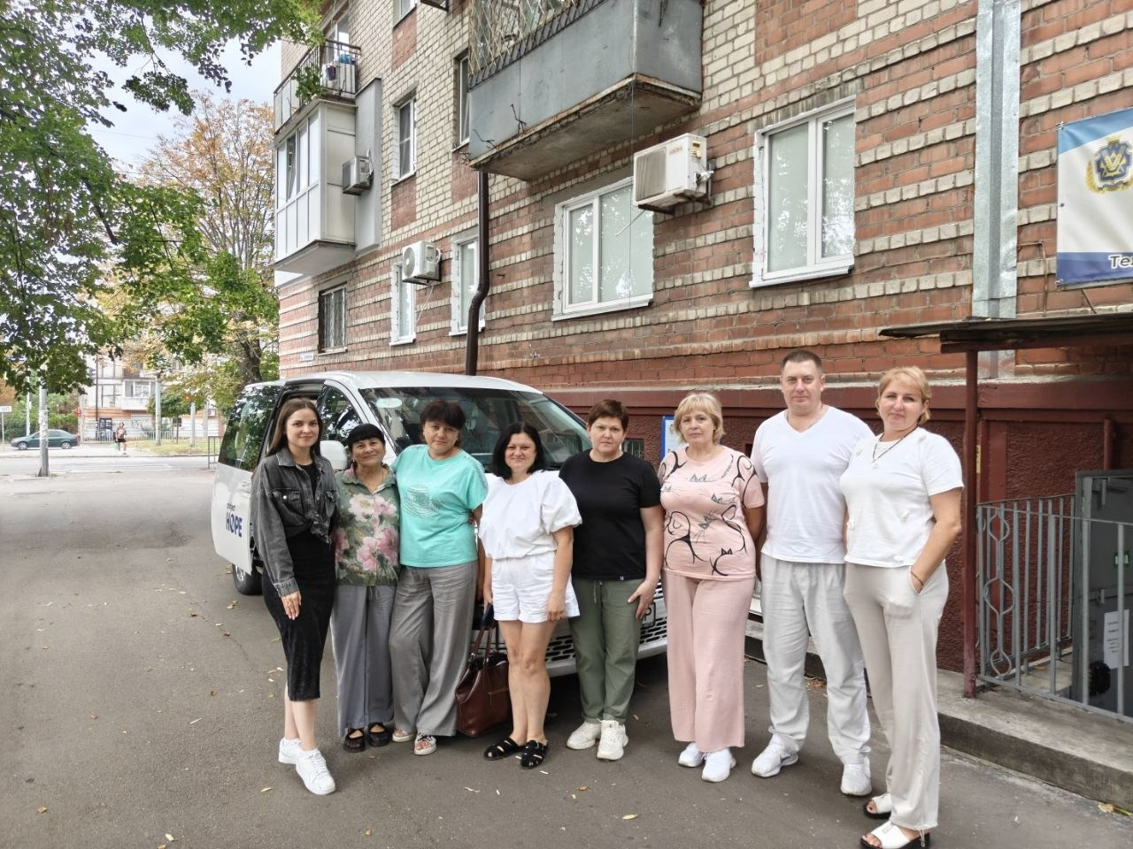 На фотографії зафіксовано колективне групове фото медичних працівників Нововоронцовської центральної районної лікарні та представників координаційного центру «Вільні разом» у Дніпрі. Вісім учасників виїзного прийому (сім жінок та один чоловік) у літньому світлому повсякденному та спортивному одязі стоять в один ряд на асфальтованій доріжці біля будівлі хабу, де відбулися 96 медичних консультацій та діагностичних обстежень для переселенців із Херсонщини, включаючи огляди гінеколога, терапевта, УЗД та флюорографію. Вони з усмішками позирують на тлі цегляної багатоповерхівки та білого мікроавтобуса виїзної медичної бригади, на борту якого нанесено маркування «HOPE», а праворуч біля входу до центру видно частину офіційної вивіски із зображенням прапора Херсонської області.