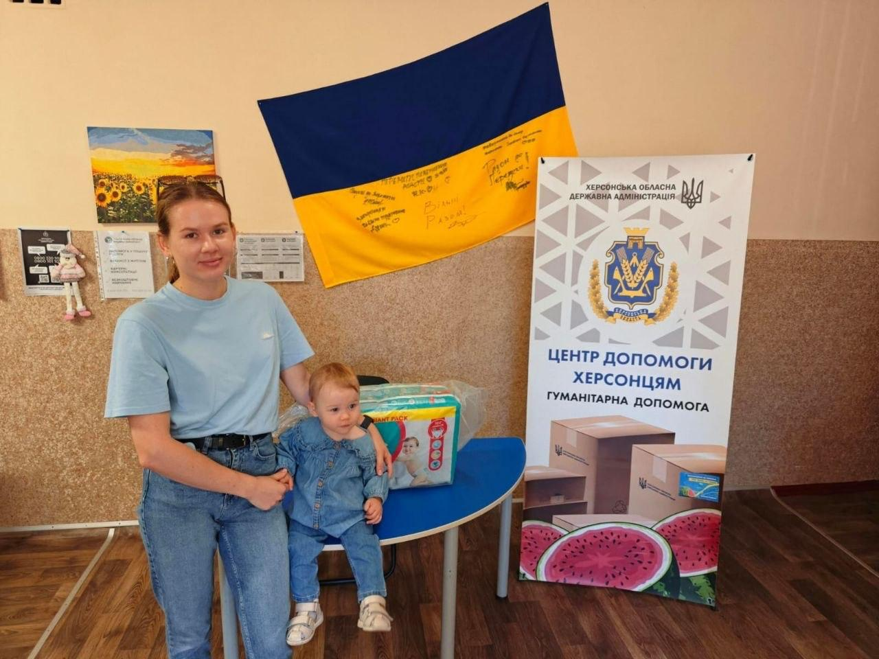 Молода жінка у блакитній футболці та джинсах отримує засоби дитячої гігієни в Хмельницькому координаційному центрі «Вільні разом». Вона стоїть поруч із маленькою дитиною в джинсовому костюмі, яка сидить на круглому столі біля великої упаковки підгузків. На стіні позаду них висить прапор України з автографами та побажаннями. Праворуч розташовано банер Херсонської ОДА з гербом області, малюнком кавунів та написом «Центр допомоги херсонцям. Гуманітарна допомога».
