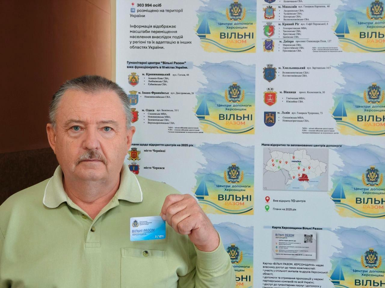 Чоловік середнього віку у світло-зеленій футболці-поло стоїть на фоні інформаційного стенда гуманітарних центрів «Вільні Разом». У лівій руці перед собою він тримає пластикову «Картку херсонця» із синьо-жовтим дизайном та написом «Вільні Разом. Херсонщина». Стенд позаду містить карту України з позначенням діючих та запланованих центрів допомоги, перелік міст (Кропивницький, Івано-Франківськ, Одеса, Миколаїв, Кривий Ріг, Дніпро, Хмельницький, Вінниця, Львів), адреси філій та назви громад Херсонської області. Також на банері розміщено логотипи «Центри допомоги Херсонцям. Вільні Разом» із малим гербом області та текстові блоки з інформацією про можливості, які надає ця картка.