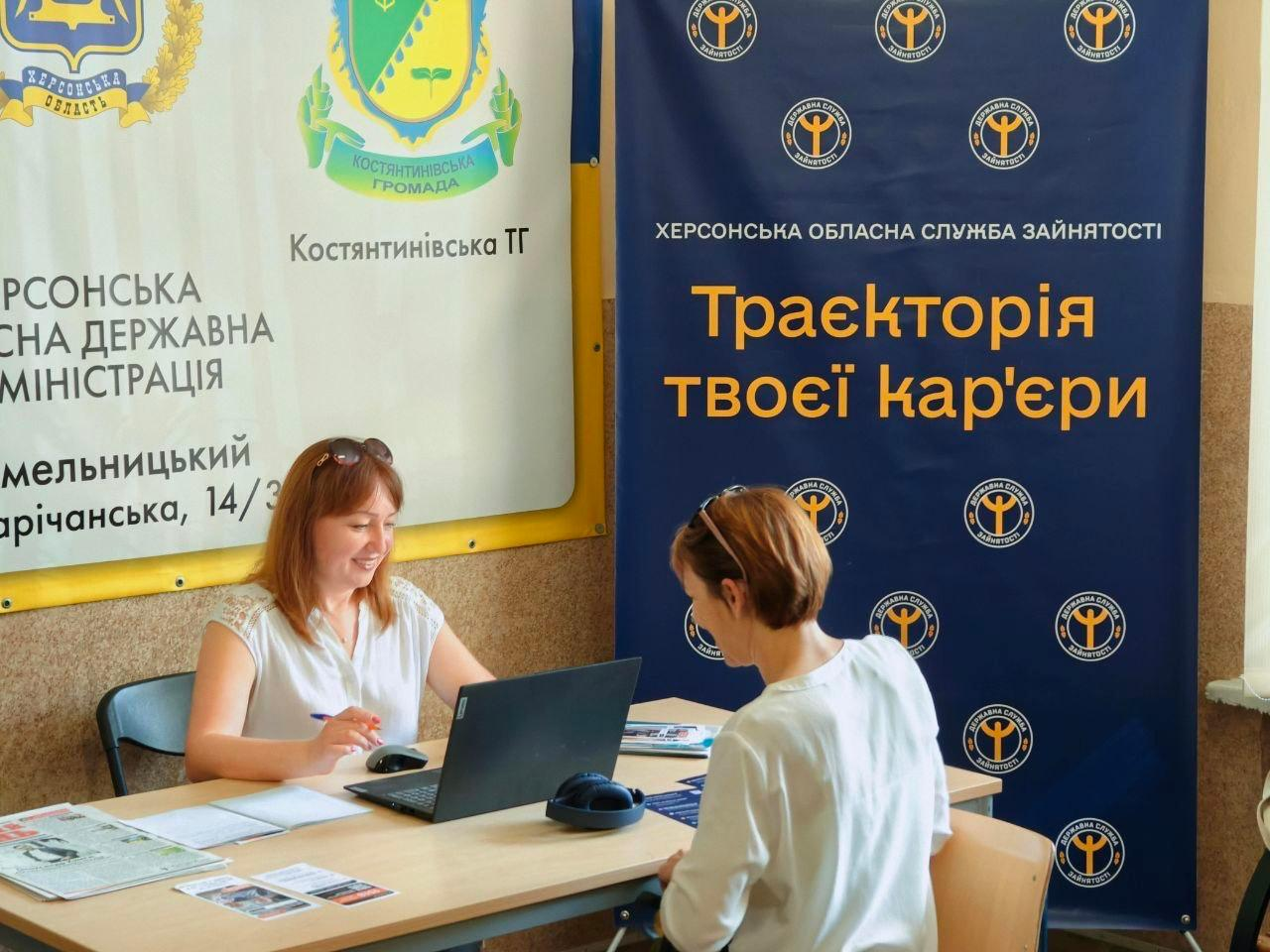 Фахівчиня Херсонського обласного центру зайнятості консультує переселенку в Хмельницькому координаційному центру «Вільні разом». Працівниця у білій блузці посміхається та тримає ручку, сидячи за робочим столом із відкритим ноутбуком і друкованими буклетами. Навпроти неї сидить відвідувачка у білій кофті. Позаду них встановлено два банери: лівий — із гербами Херсонської області та Костянтинівської ТГ, правий — темно-синій банер Херсонської обласної служби зайнятості з логотипами та помаранчевим гаслом «Траєкторія твоєї кар'єри».